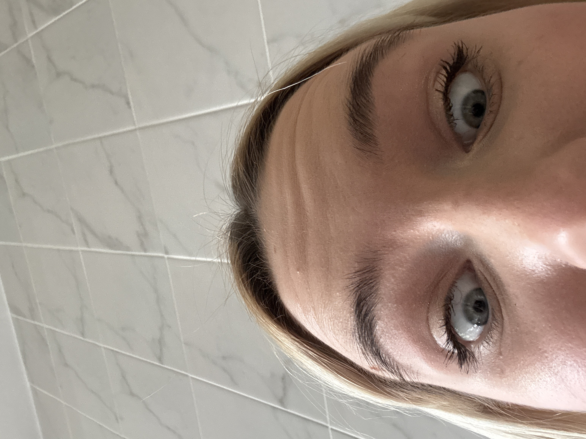 A close-up, tilted perspective of a person's face, focusing on the eyes, eyebrows, forehead, and hairline. The eyes are light-colored, with visible eyelashes and mascara. The eyebrows are dark and thick. The forehead shows some skin texture and minor wrinkles. Blonde hair is visible near the hairline. The background shows a tiled wall with a marble pattern. The image does not contain information related to the city of Saint Petersburg, Russia.