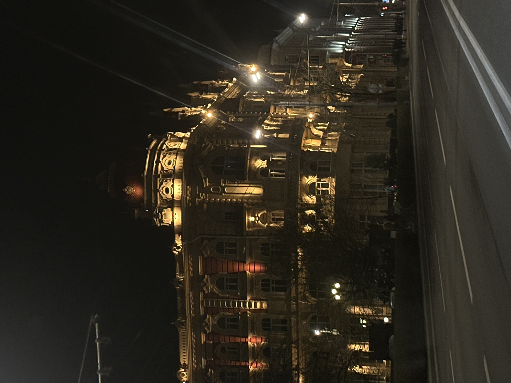 The image shows a building at night, likely the Taj Mahal Palace hotel in Mumbai, India, not Rostov-on-Don, Russia. The hotel's iconic dome is partially visible on the left. The building is illuminated with lights that highlight its architectural details. To the right of the hotel is a road. There is very little light. The sky is dark.