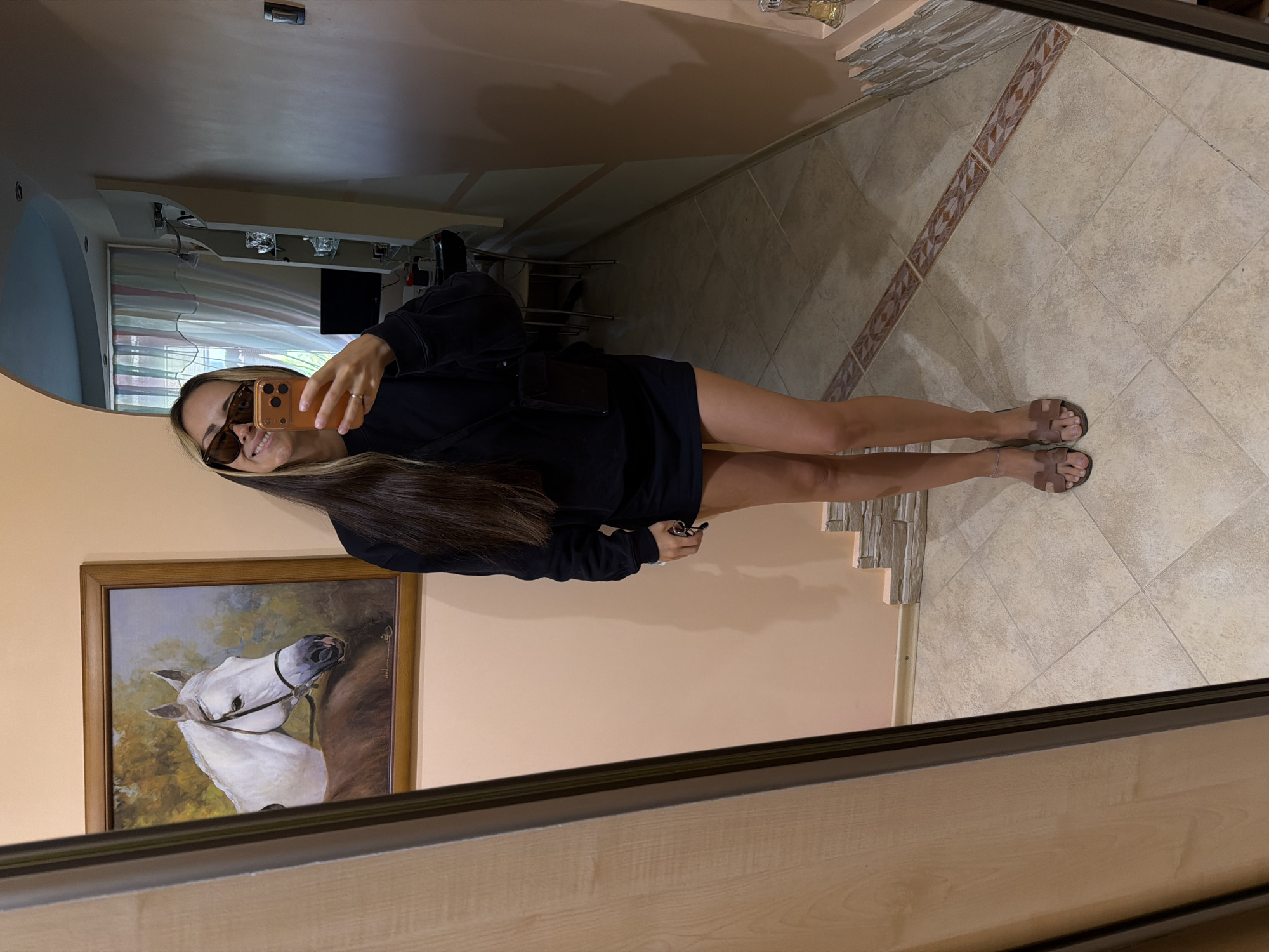 A young woman is taking a mirror selfie in what appears to be a hallway. She is wearing sunglasses, a black long-sleeved shirt, a black skirt, and brown sandals. She has long dark hair with blonde streaks. She is holding an orange smartphone in her right hand, and her left hand is holding her keys.
In the background, there is a framed painting of a white horse on the wall. There is also a door, some shelving with decorative items, and a computer monitor. The flooring is tiled. The lighting appears to be artificial. The overall impression is an indoor residential setting. There is no clear indication of the location in the city of Kamyshin, Russia.
