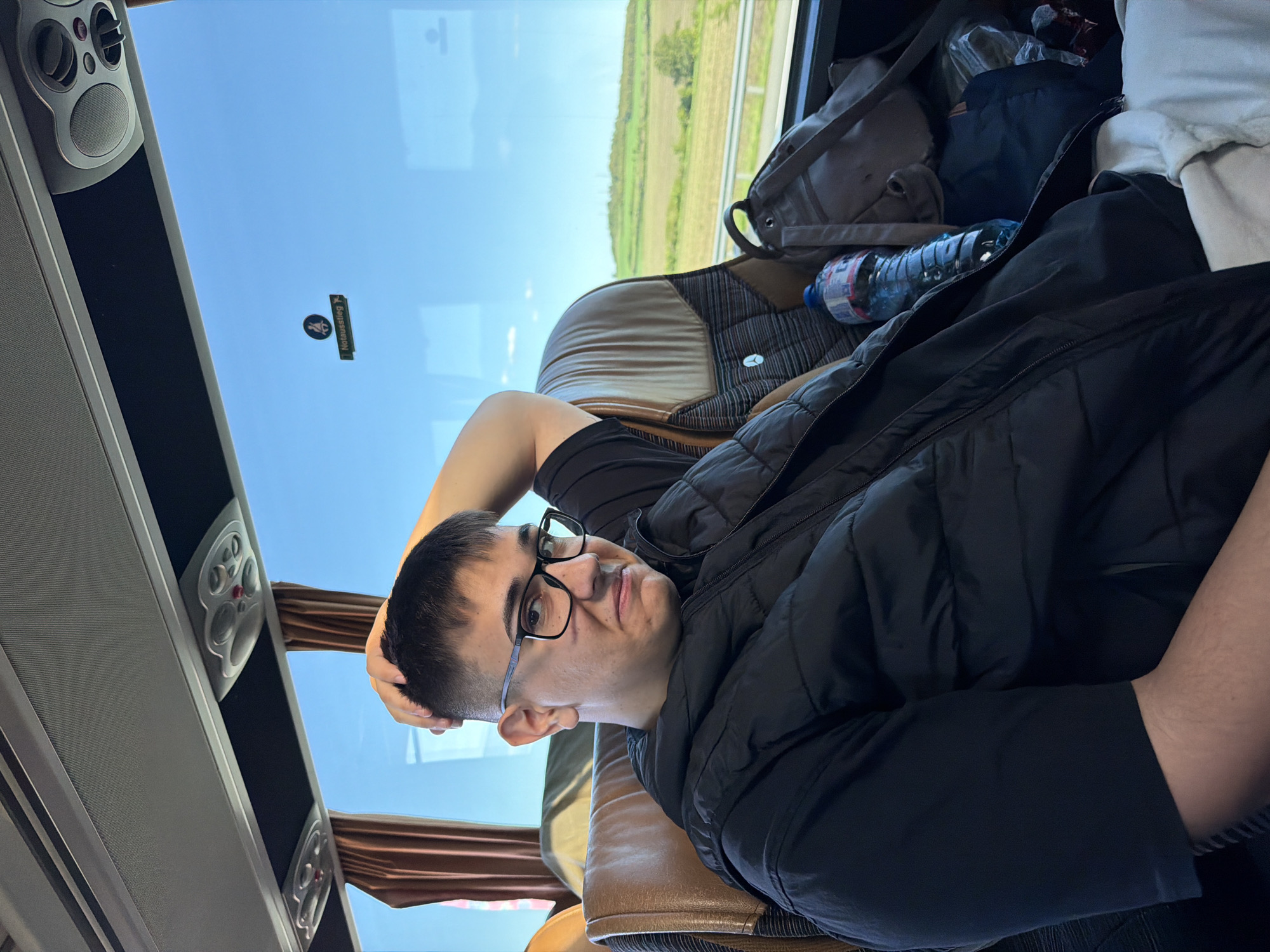 A person is lying on a bus seat, with their head resting on their hand. They are wearing glasses and a black jacket. A water bottle and bags are next to them. The bus window shows a view of fields and trees. The interior of the bus features speakers and overhead compartments. The image provides no information to confirm the location is Yazdach, Bulgaria.