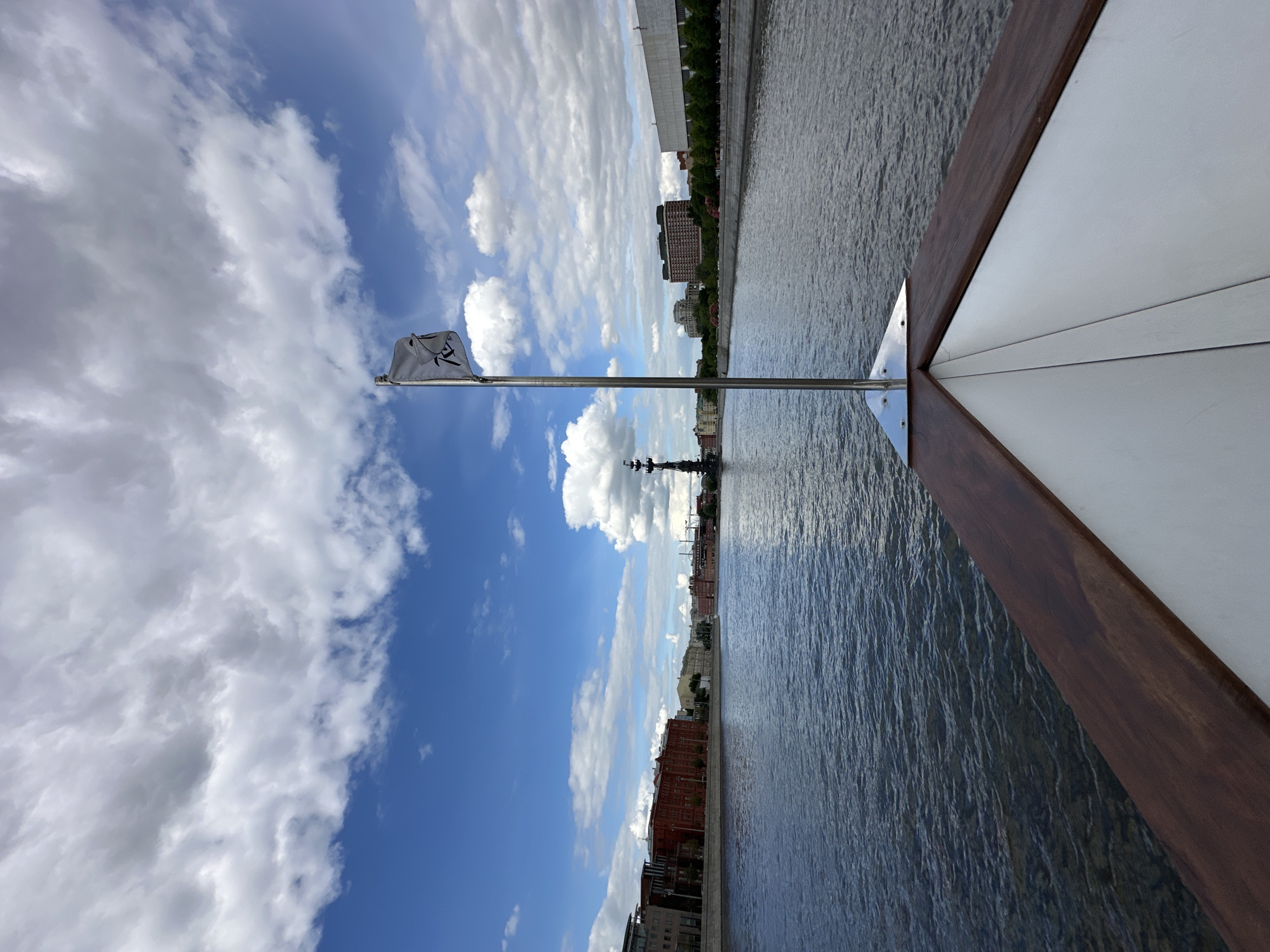 The image shows a view from a boat on a body of water, likely the Moskva River. The angle suggests the photograph was taken from the front of the boat. The sky is blue with scattered clouds. A flag with an emblem on it is affixed to the front of the boat. The cityscape on the riverbank includes buildings and structures characteristic of Moscow, Russia. The Peter the Great statue is visible.