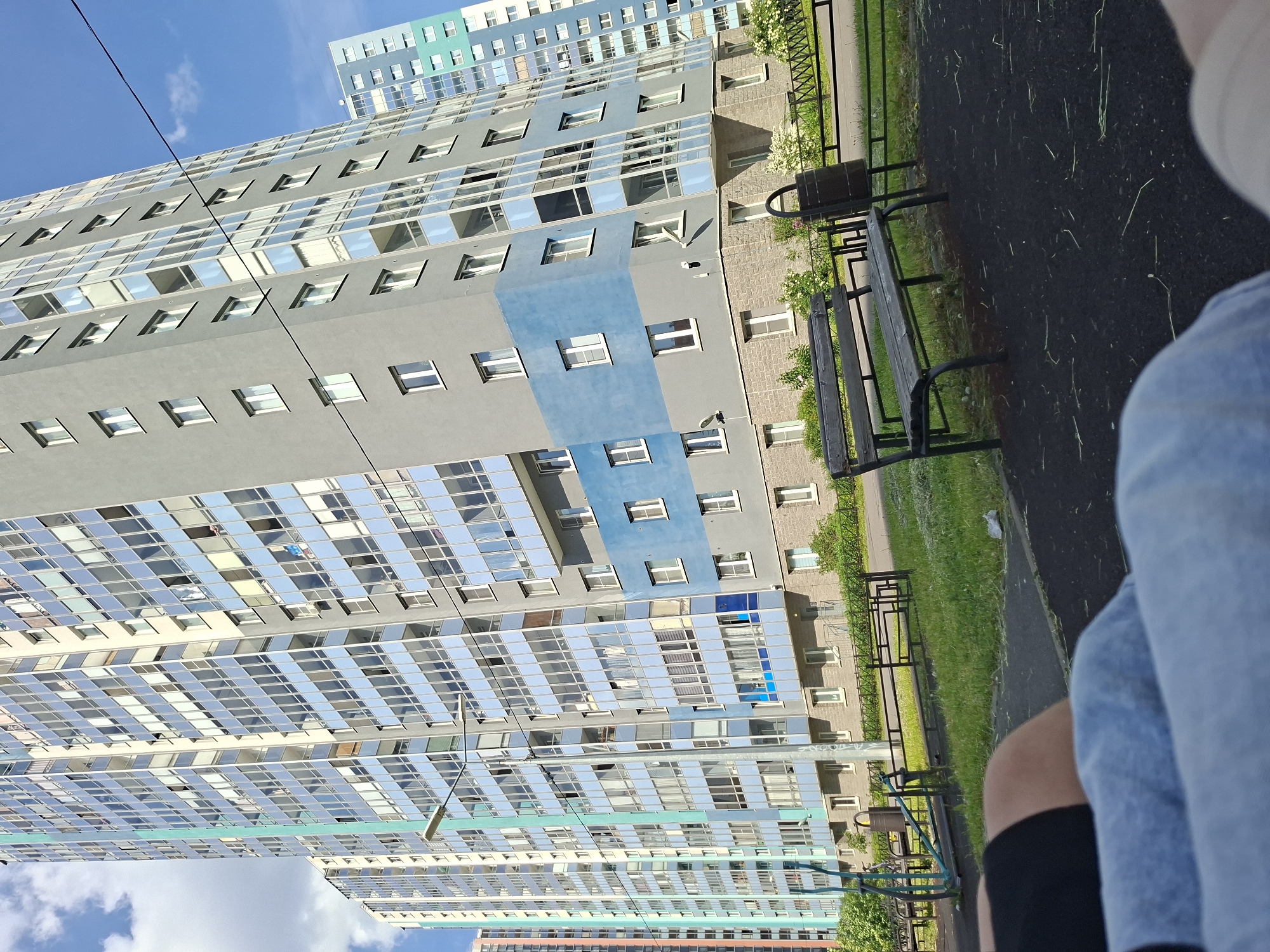 This rotated image captures a sunny urban landscape dominated by modern apartment buildings, viewed from a low perspective. In the immediate foreground, to the right, are the legs of a person, one clad in light blue denim and the other showing bare skin above dark fabric, suggesting they are seated on or near the ground.
The scene features a dark paved surface with sparse blades of grass, which transitions into a neat strip of green lawn. A dark wooden park bench with a black metal frame is positioned on the grassy area, next to a matching black trash can. A low, dark metal fence or railing runs along the edge of the green space, separating it from the buildings.
The background is filled with tall, multi-story residential buildings. The most prominent building displays a contemporary design, primarily gray, interspersed with vertical and horizontal sections of light blue. Its facade is punctuated by numerous windows, many of which are highly reflective, mirroring the bright blue sky and clouds. Further back, additional buildings with similar modern architecture feature lighter colors and hints of teal. The overall impression is of a vibrant, recently developed residential area.
The sky is a clear, bright blue with a few wispy white clouds, indicating sunny weather. Strong shadows are cast by the buildings and foreground objects, suggesting it is daytime, likely mid-morning or afternoon. There is no visible text in the image. The setting is identified as Saint Petersburg, Russia, consistent with the modern residential architecture.