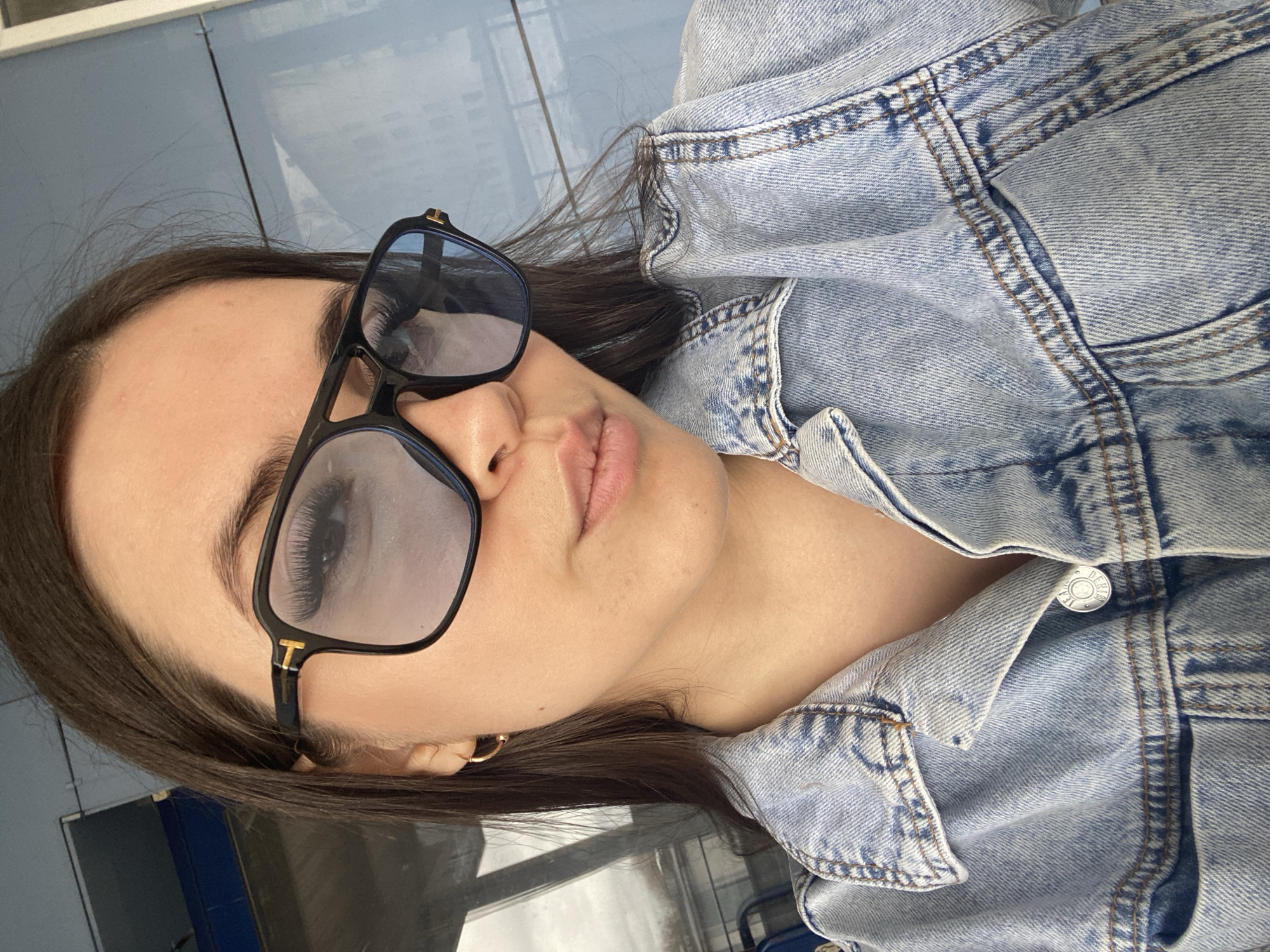 A woman with long brown hair is wearing sunglasses and a denim jacket. She is situated indoors with blue tiled flooring. The location is the city of Chaikovsky, Russia.