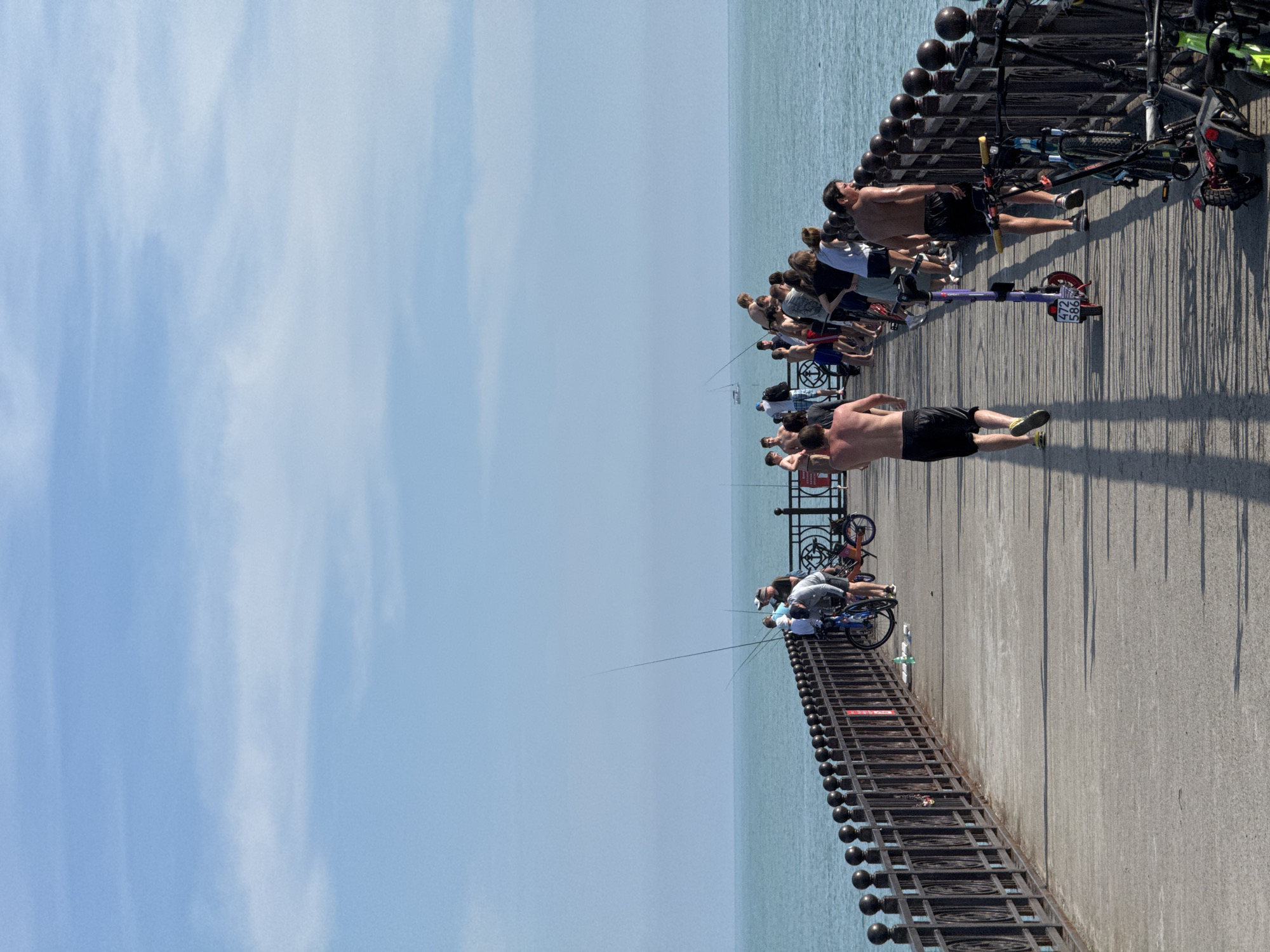 The image shows a concrete pier extending into the Black Sea in Sirius, Russia, under a bright blue sky with sparse, wispy clouds. The pier is lined on both sides by a dark, ornate metal railing. Numerous individuals are present on the pier. On the left side, several people are standing near the railing, some actively fishing with rods extending over the calm, turquoise water. A person is crouched beside a bicycle on the left, and a green bottle rests on the pier surface. On the right side of the pier, a larger group of people is visible. Two shirtless men in dark shorts are walking away from the viewer in the foreground, and another shirtless man in dark shorts is running down the pier, casting a long shadow. Multiple bicycles are parked along the railing on the right, along with a purple electric scooter displaying license plate "472 586". A small white boat is discernible in the distant sea. Long, distinct shadows indicate strong directional sunlight.