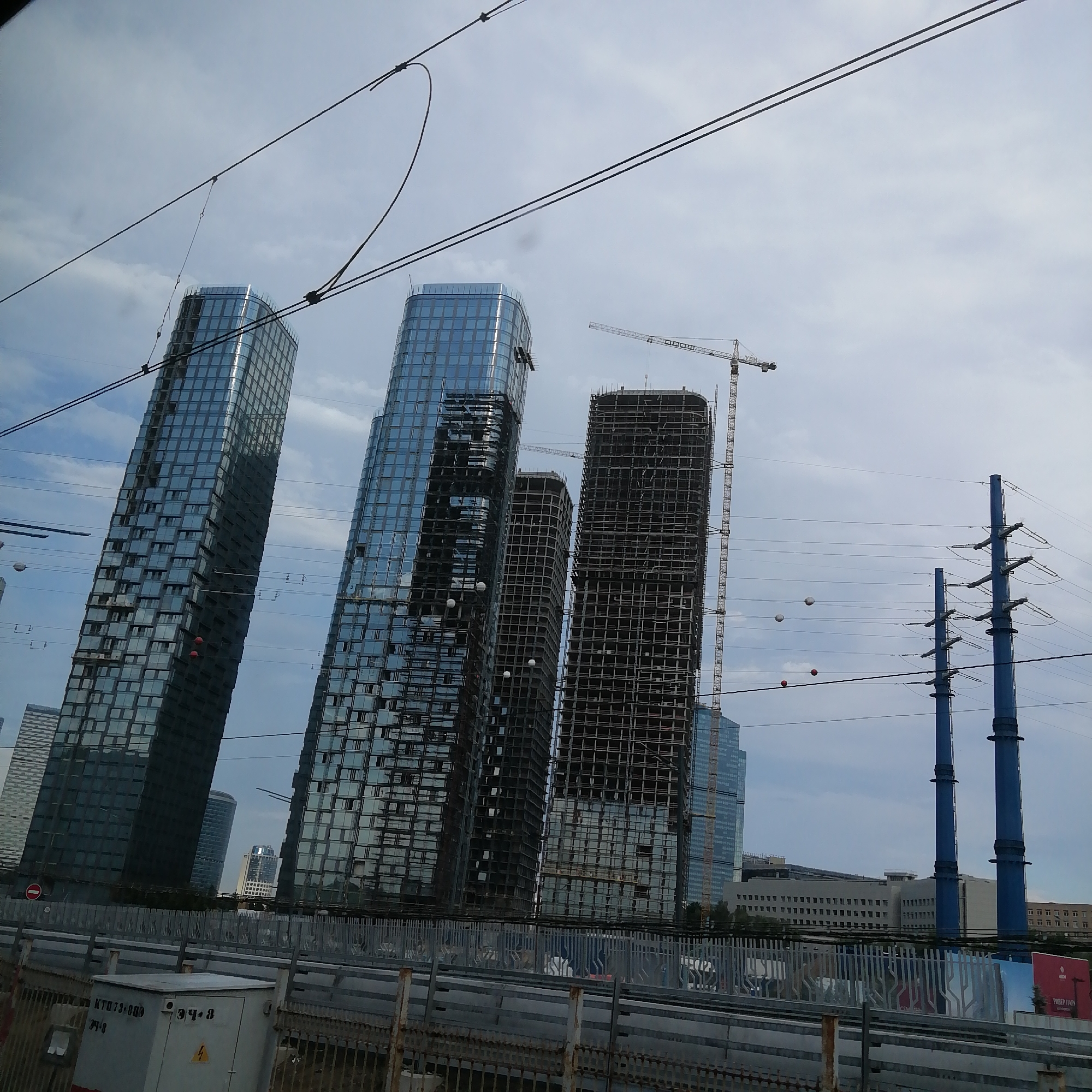 The image shows a view of modern skyscrapers under construction in an urban environment, possibly Moscow City, against a cloudy sky. In the foreground, there is a barrier or fence with some electrical equipment to the left. Several tall buildings are visible, with two particularly prominent ones featuring glass facades. One of these is mostly completed, while the other appears to be in an earlier stage of construction, with its skeletal framework exposed. To the right, a construction crane is visible, indicating ongoing building work. Further in the distance, other buildings, including one with a more rounded shape and another that appears completed with a distinct architectural design, can be seen. Utility poles with power lines run across the scene, adding to the industrial and urban feel. The overall impression is one of a dynamic, developing cityscape.