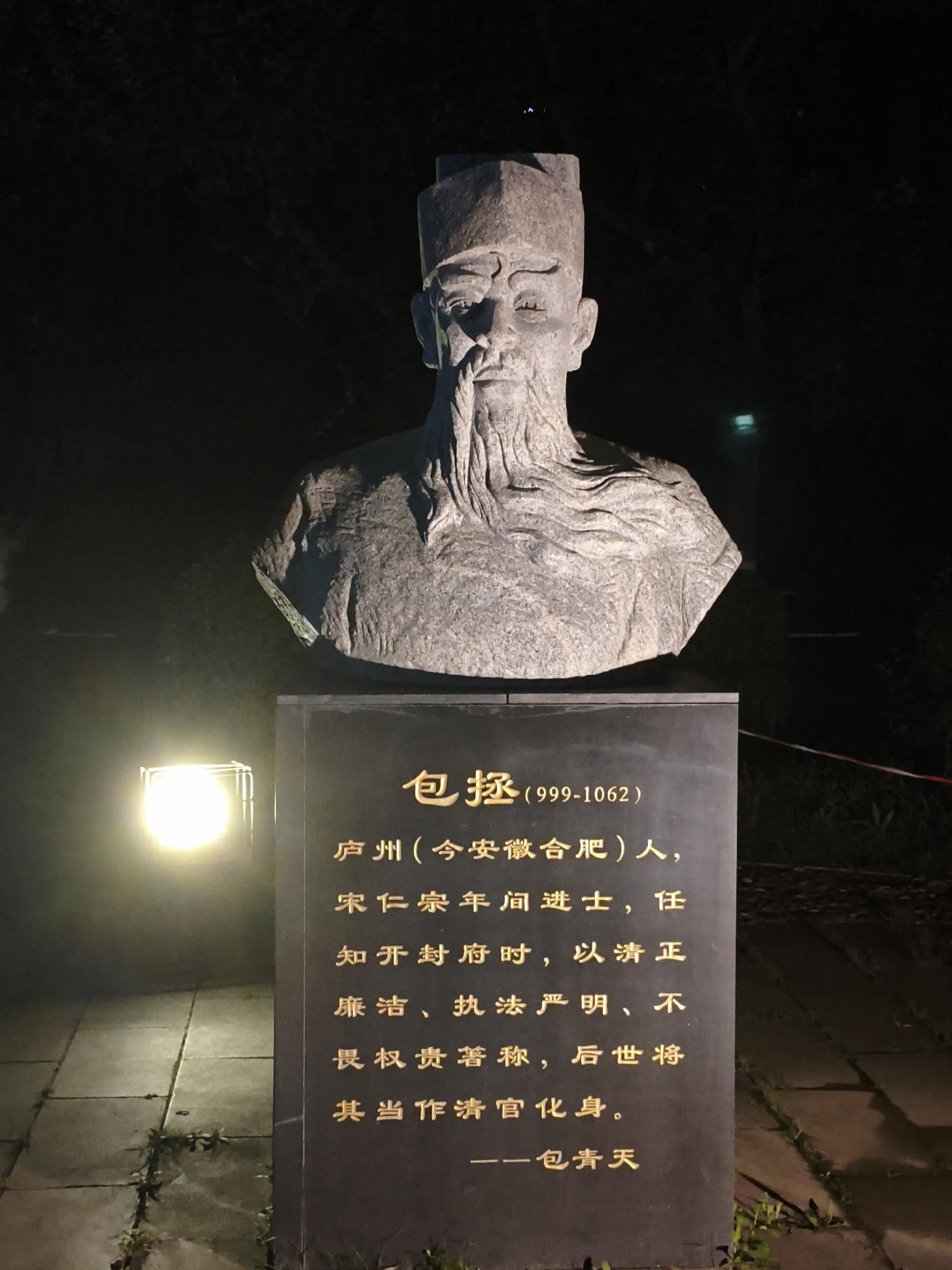A stone bust of a man with a hat and beard stands on a dark stone monument at night. The monument has gold lettering in Chinese. The bust faces forward, with its head slightly tilted. The monument is lit from the left by a bright light source, casting shadows on the ground. The background is dark, with some faint lights and foliage visible. The scene appears to be outdoors, possibly in a park or a public square. The monument is located in Dongxing Street, China.