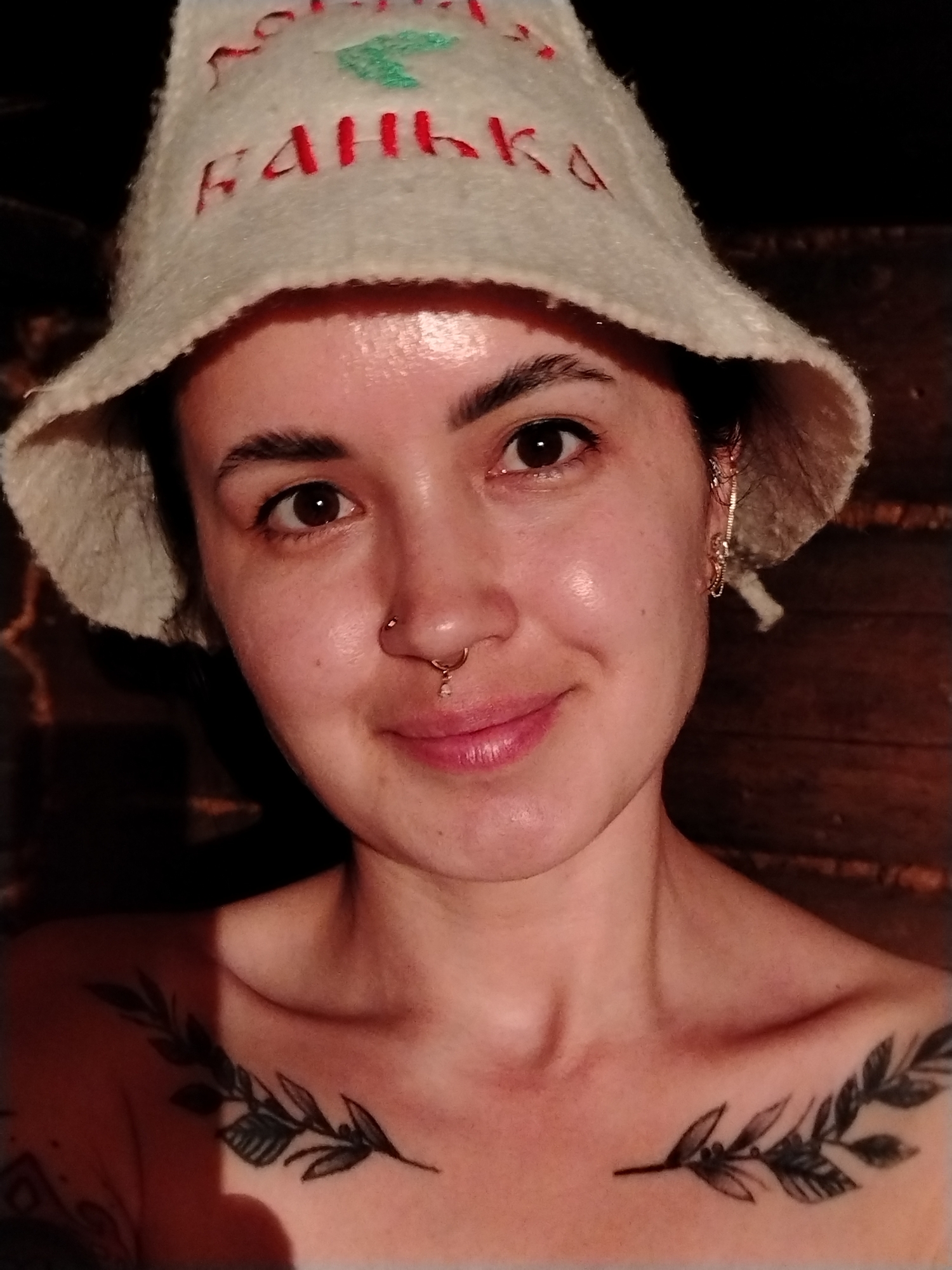 A close-up shot shows a woman with dark hair wearing a traditional felt sauna hat with the word "БАНЬКА" (Banya) stitched on it in red and green. She has a nose piercing and gold earrings. A tattoo of two leafy branches is visible on her upper chest. The background appears to be a dark, wooden interior, possibly a sauna. The image was taken in the city of Гожан, Russia.