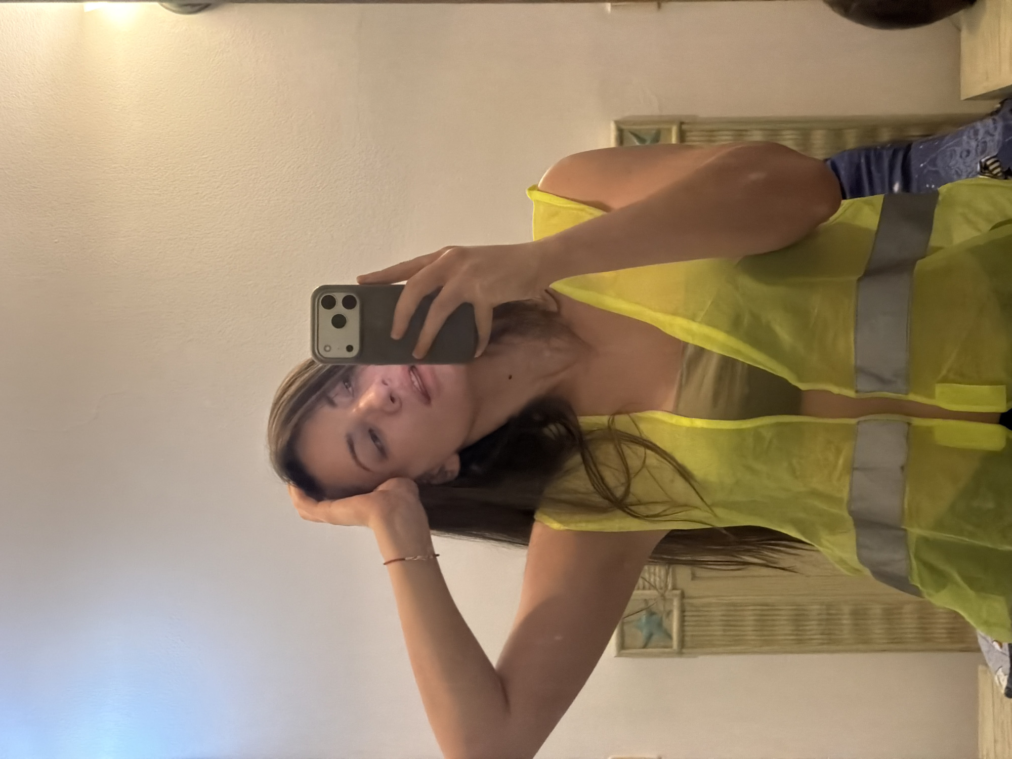 A woman stands in front of a mirror, taking a selfie. She is wearing a neon yellow safety vest over a light brown top. Her left arm is raised, with her hand touching her head. She has long, dark hair. The background appears to be a wall with some decorative elements and a light fixture. There is no indication of the city of Sharm El Sheikh, Egypt.