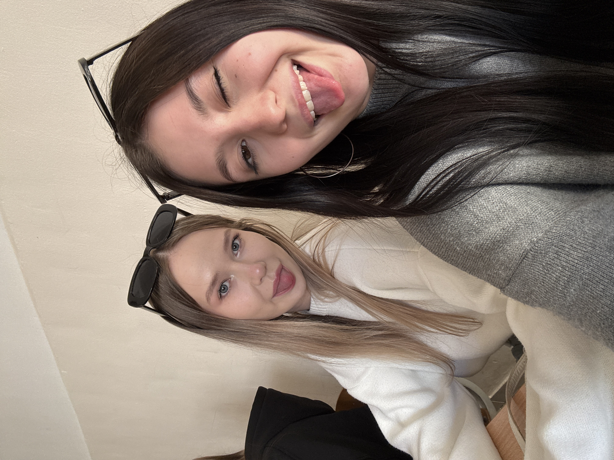 Two young women are posing for a selfie. The woman at the top has dark hair and is wearing glasses and a gray sweater. She is winking and sticking her tongue out. The woman at the bottom has blonde hair and is wearing sunglasses on her head and a white sweater. She is also sticking her tongue out. The background is a light-colored wall. The city of Chaikovsky, Russia, is not identifiable in the image.