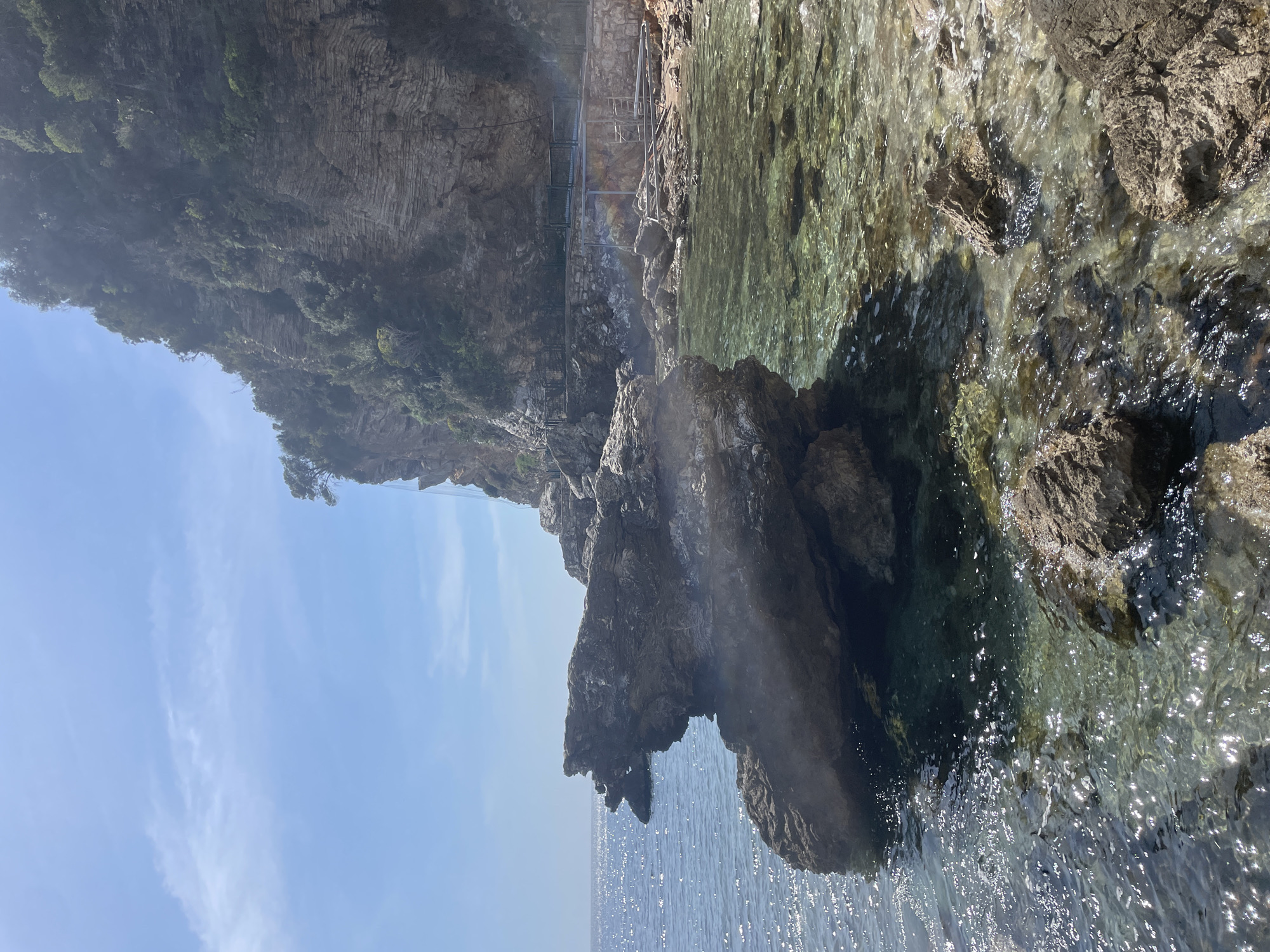 The scene depicts a rugged coastal area along the Adriatic Sea in Budva, Montenegro, likely situated near the pathway to Mogren Beach. The foreground and midground feature clear, shallow seawater with a discernible turquoise tint, revealing a rocky seabed. Sunlight glints on the water's surface, indicating gentle ripples. A large, dark, irregularly shaped rock formation protrudes from the left-center into the water. To the right, a steep, layered rock cliff face, densely covered with green trees and shrubs, rises vertically. Along the base of the cliff, a concrete pathway is visible, supported by a metal framework and featuring dark metal railings. This path includes an elevated section with integrated steps descending towards the rocky shoreline. Additional metal structures, consistent with a ladder or support, are positioned on the rocks adjacent to the path. The sky above is clear and blue with minimal cloud cover. No individuals are present in the depicted environment.