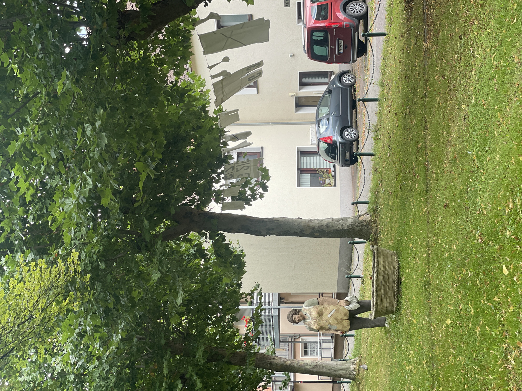 In Salzkotten, Germany, a statue of a woman stands on a green lawn in front of a light-colored building. The statue depicts a woman wearing a patterned vest over a green shirt and dark pants, holding a white cloth in her left hand and what appears to be a white handkerchief or napkin in her right. In front of the statue is a large, woven basket. To the right of the statue, two cars are parked: a dark gray hatchback and a red van. Laundry, consisting of large white fabric pieces, is hung on a clothesline strung between trees, with some pieces displaying dark lettering, including "C3". The scene is characterized by lush green trees with dense foliage, casting shadows on the grass. A low, dark metal chain fence with green posts separates the grassy area from the paved area where the cars are parked. The building behind the statue and laundry has windows and balconies, suggesting it is a residential or commercial structure. The overall impression is a daytime scene in a semi-urban environment, possibly a courtyard or public space within Salzkotten.