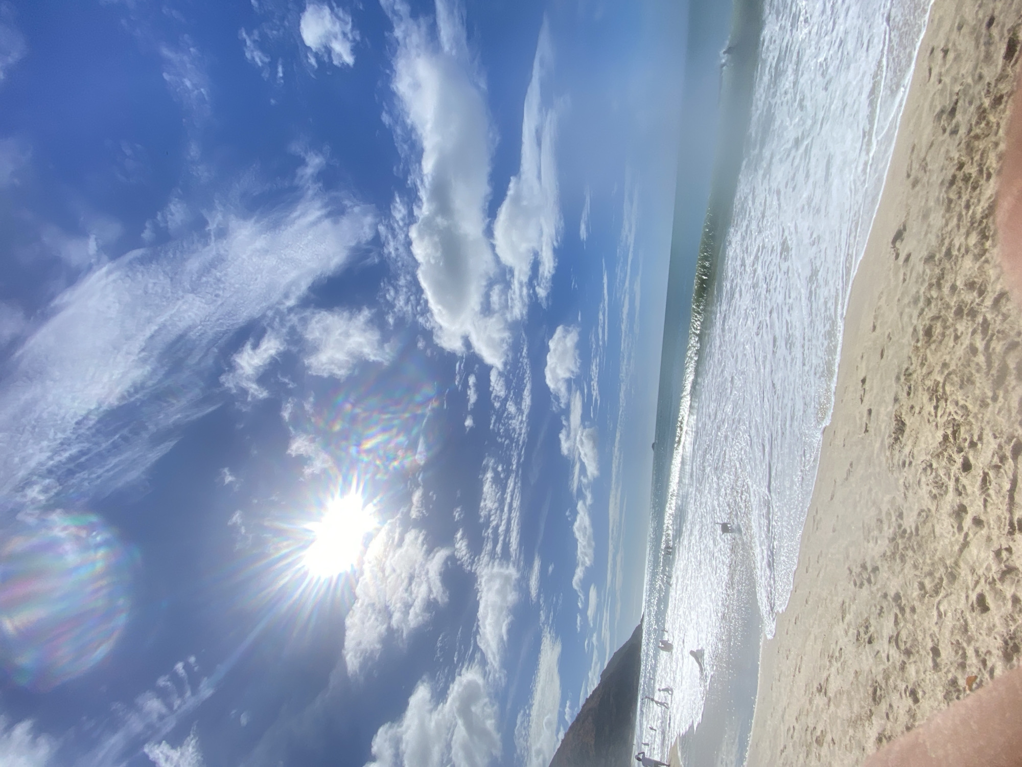 The image depicts a beach scene in Pedro González, Venezuela, taken at an oblique angle. A bright sun is visible in a blue sky filled with scattered white clouds. The beach is sandy with visible footprints, and the shoreline is marked by gentle waves. Several people are present on the beach, some near the water's edge. A hill or rock formation rises at one end of the beach.