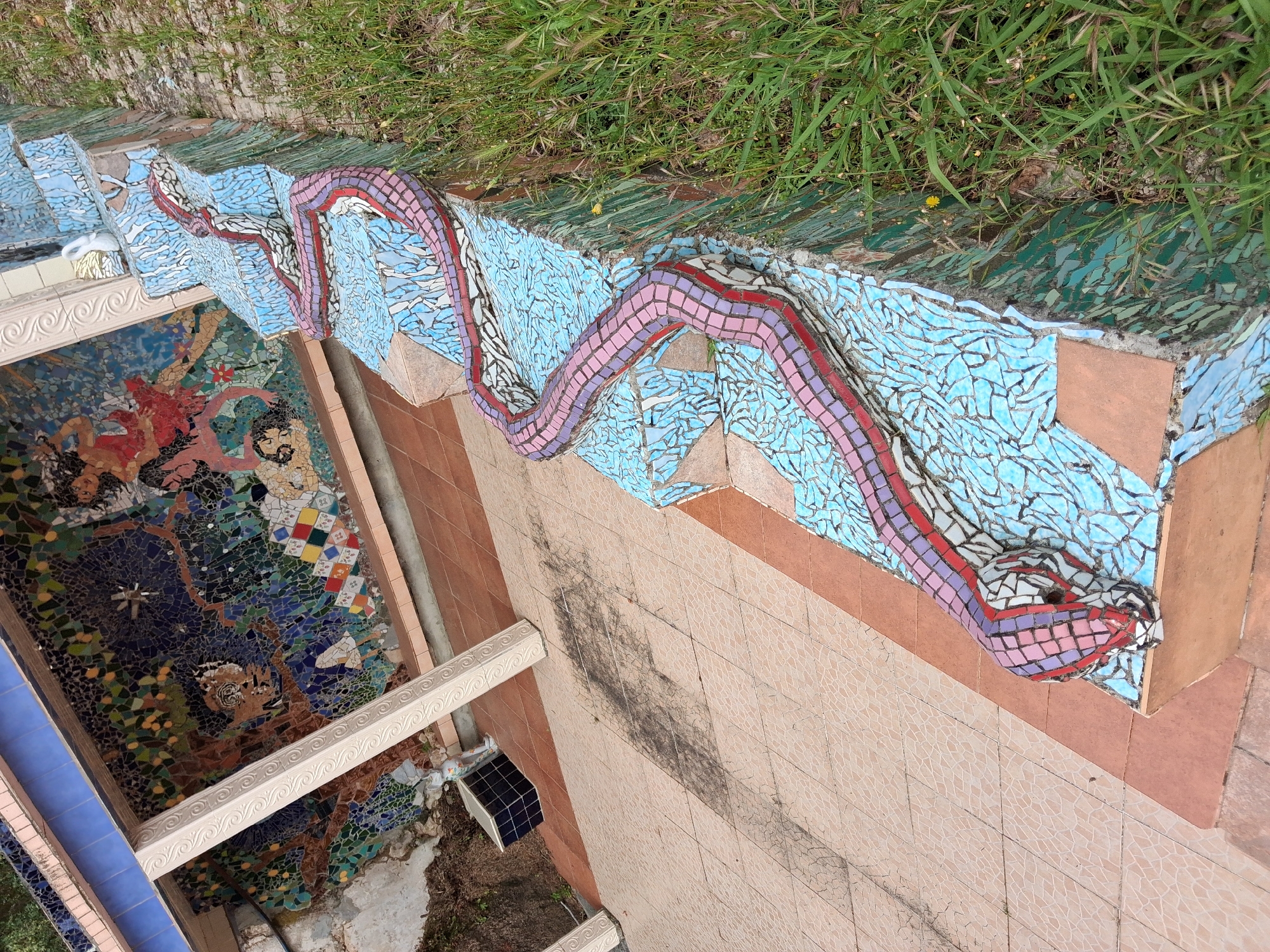 The image depicts a mosaic artwork in Soure, Portugal. The scene includes a descending series of steps decorated with a mosaic depicting a serpentine creature with light blue, red, purple, and white tiles. Above the stairs is a patch of green grass. To the left, a wall covered in a mosaic mural is visible. Adjacent to the stairs is a tiled wall with a light peach color, showing dark stains. A portion of a white ornate rail runs along the edge of the mosaic mural. A dark colored tile cube is visible, in the ground to the left of the mosaic wall. The overall composition exhibits a unique combination of art, architecture, and natural elements.