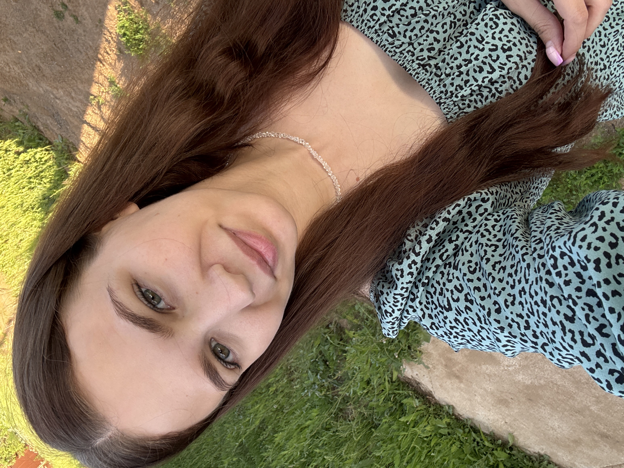 A young woman with long, brown hair is captured in a selfie outdoors. She is wearing a light blue top with a black leopard print and a delicate, clear beaded necklace. Her green eyes are looking directly at the camera, and she has a slight smile. The background suggests an outdoor setting with green grass and a dirt path, indicative of a suburban or rural area. The lighting suggests it is daytime, possibly late afternoon due to the warm tones. The image is a close-up, focusing on the woman's face and upper body. There is no explicit indication of the specific location within Nizhnyaya Gar', Russia, but the environment is consistent with a park or backyard in such a region.