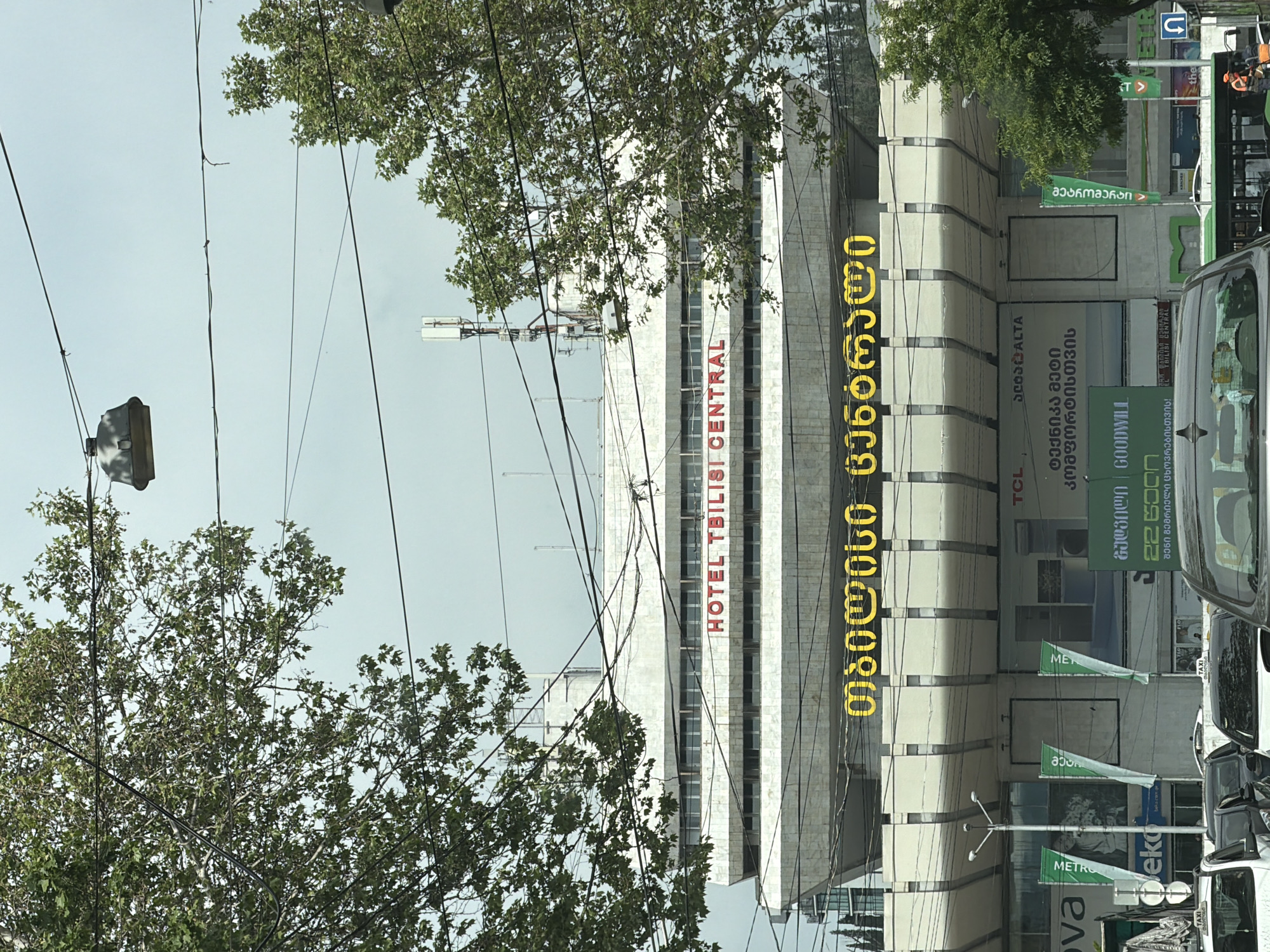 The image shows a street view in Tbilisi, Georgia, featuring the Hotel Tbilisi Central. The hotel's name is displayed in both English and Georgian on the building's facade. There are trees and power lines in the foreground, and various signs and advertisements can be seen along the street. A vehicle is partially visible on the right side of the frame, suggesting a bustling city environment.