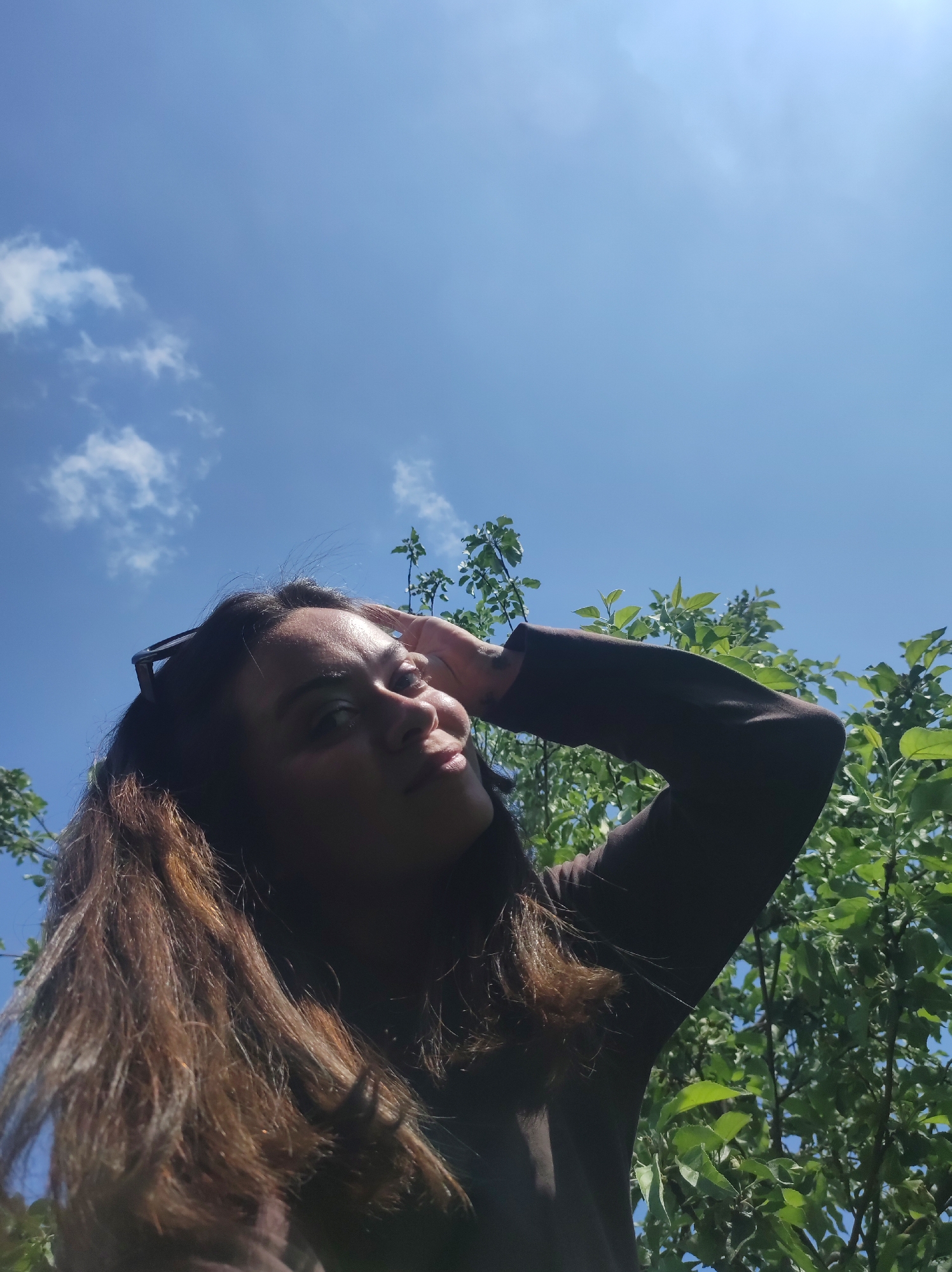 The image features a low-angle shot of a woman outdoors in Chaikovsky, Russia, on a bright, sunny day. The sky, a vibrant blue with scattered white cumulus clouds, dominates the upper half of the frame. Lush green foliage from trees or bushes is visible on the left and right sides, and also in the mid-ground behind the woman, suggesting a natural setting like a park or garden.
The woman, with long, dark brown hair, is looking slightly upwards and to her right. Her face is partially shadowed, but her features are discernible, showing a calm expression with a subtle, slight smile. She has sunglasses perched on top of her head. Her right hand is raised, touching her forehead or hair, possibly shielding her eyes from the bright sunlight or as a casual pose. She is wearing a dark, long-sleeved top. The strong sunlight and clear sky indicate it is daytime, likely mid-day. The overall impression is one of relaxed enjoyment in the outdoors. No text is visible in the image.