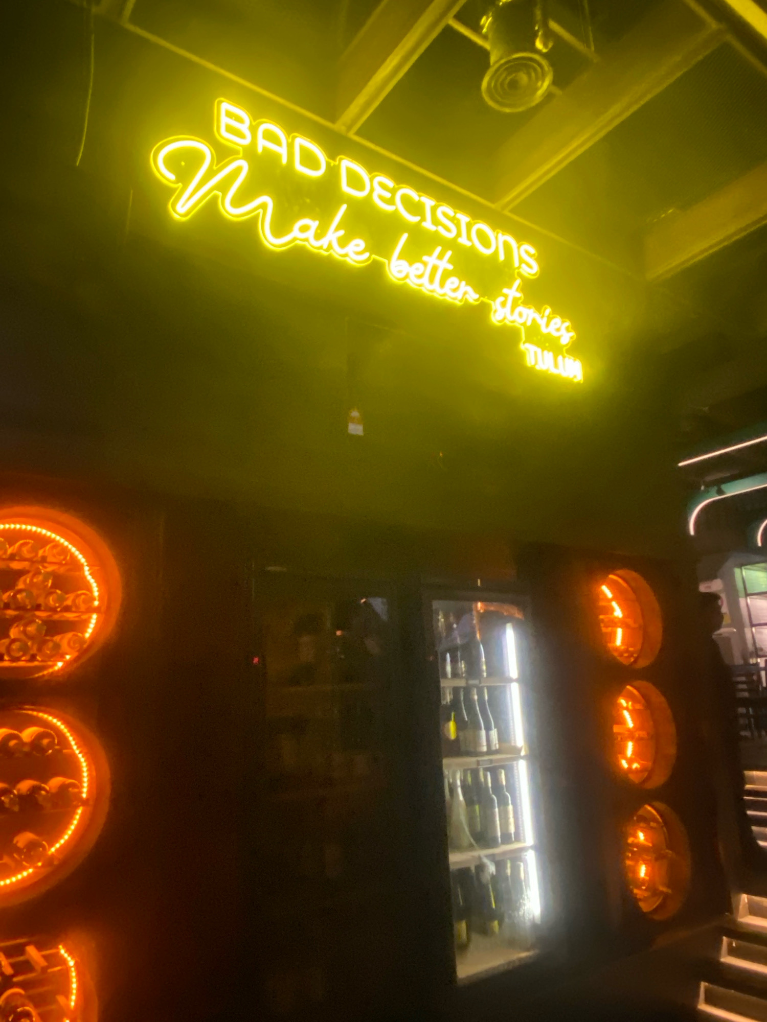 In a dimly lit establishment, a neon sign glows bright yellow, displaying the text "BAD DECISIONS Make better stories" above a smaller line of text that reads "TULUM". The sign is mounted against a dark ceiling with visible beams and a spotlight. Below the sign, a dark wall features a glass-fronted refrigerator filled with bottles, illuminated by a bright vertical light. To the left of the refrigerator are three circular display units, each containing bottles and outlined with orange lights. To the right of the refrigerator, there are visible steps and a silhouette of a person. The overall ambiance suggests a bar or restaurant setting, possibly with a Tulum-themed decor, located in Kuala Lumpur, Malaysia.