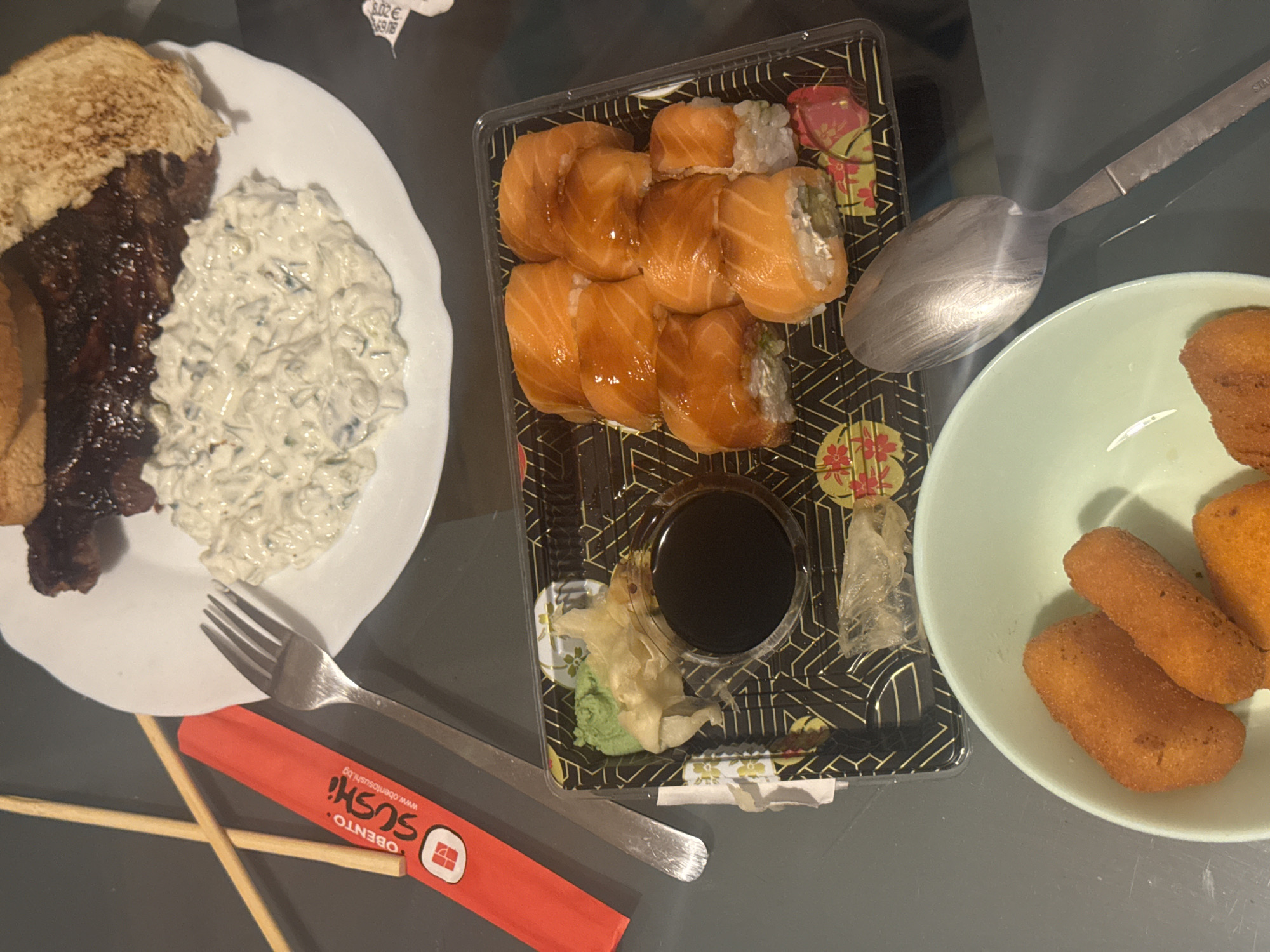 The image shows a tabletop view of a meal consisting of three plates/containers: a white plate with a meat dish, accompanied by a light-colored, creamy salad; a black plastic sushi container with salmon sushi, soy sauce, wasabi, and ginger; and a light green bowl containing fried croquettes. There are eating utensils including a fork, a spoon, and chopsticks in a red wrapper. The sushi container has the logo of "OBENTO SUSHI" and the website "www.obentosushi.bg," which indicates the meal is from Sofia, Bulgaria, as this company is based in Sofia. The setting is indoors, likely in a restaurant or home.