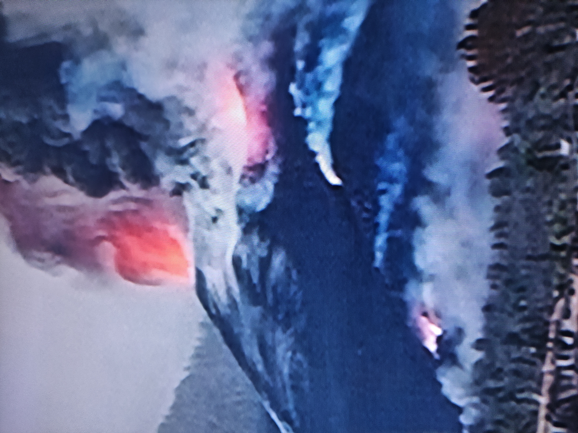 The image depicts a dramatic natural phenomenon characterized by intense fire and vast plumes of smoke. A large, dark, and rugged landmass, resembling a mountain or steep hillside, occupies the central and right areas of the frame. Bright, incandescent red and orange glows are visible at multiple points on this dark terrain, strongly suggesting either a volcanic eruption or a large-scale wildfire.
Prominent fiery hotspots appear on the left side of the landmass, where a significant glowing red area is seen, and also further down on the right. Massive plumes of smoke, varying in color from dark gray to lighter blue-gray and white, billow upwards and spread across the sky, partially obscuring the background. The interaction of the bright flames with the smoke creates dynamic lighting effects, with some smoke illuminated from within by the intense heat.
The overall lighting conditions, with the vivid glows contrasting against the darker landscape and smoke-filled atmosphere, suggest a low-light setting such as night, dusk, or dawn. The scene conveys a powerful and potentially destructive event. No people, discernible man-made objects, or visible text are present within the image. The image quality, characterized by visible pixel grids or scan lines, indicates that it is likely a photograph taken of a screen displaying this event. The provided context places this event in Pavlodar, Kazakhstan.