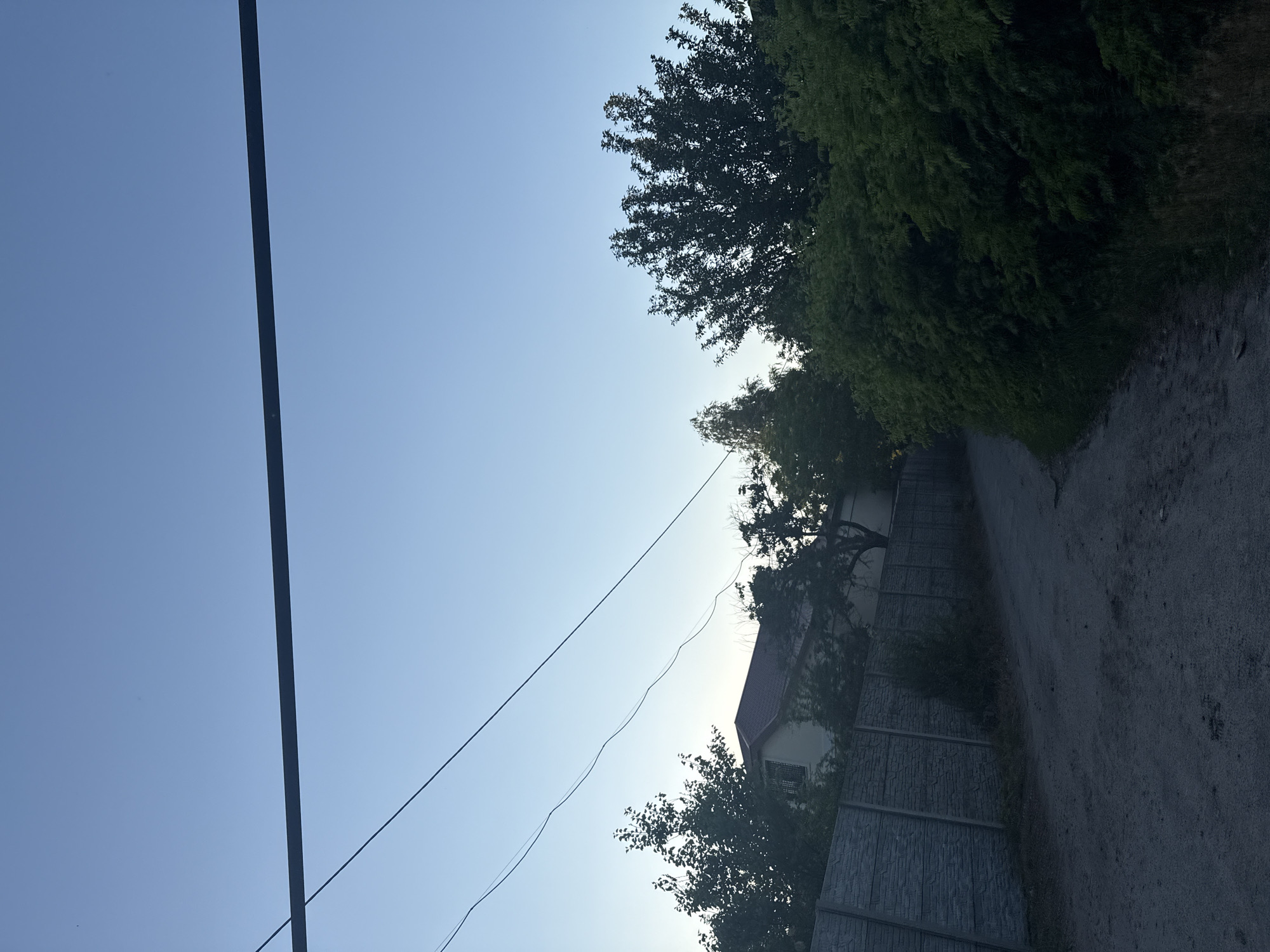 The image depicts a rural road in Obukhivka, Ukraine, under a clear blue sky. The road, made of dirt and gravel, curves to the right. On the left side of the road, a textured concrete fence runs along the property line. Behind the fence, a light-colored house with a reddish-brown roof is partially visible, surrounded by lush green trees. To the right of the road, dense green bushes and trees form a thick hedge. Two dark power lines stretch across the upper portion of the image, suspended from poles that are not visible. The scene is illuminated by natural daylight, suggesting it was taken during the daytime. No people are present in the image.