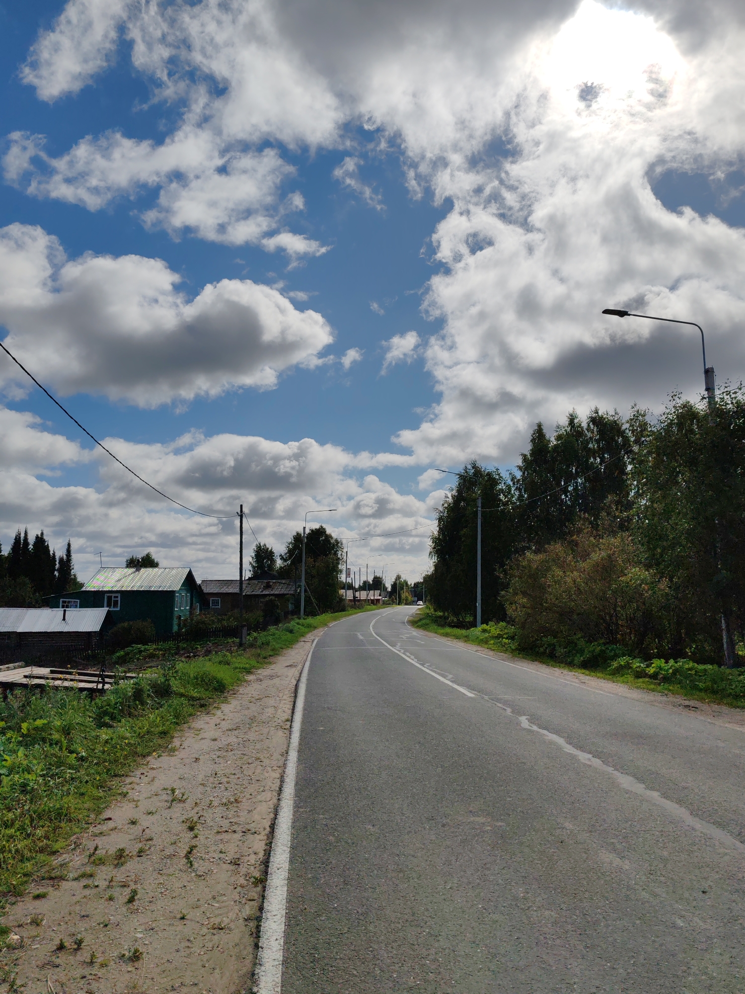 The image depicts a rural road in Чухлэм, Russia, on a partly cloudy day. The road curves to the left in the distance, flanked by a dirt and grassy shoulder on the left and trees and vegetation on the right. Several wooden houses with pitched roofs are visible on the left side of the road, one painted green and others in muted tones. Power lines stretch across the sky. A street lamp stands on the right side of the road, near a cluster of trees. The sun is visible through the clouds in the upper right portion of the image.
