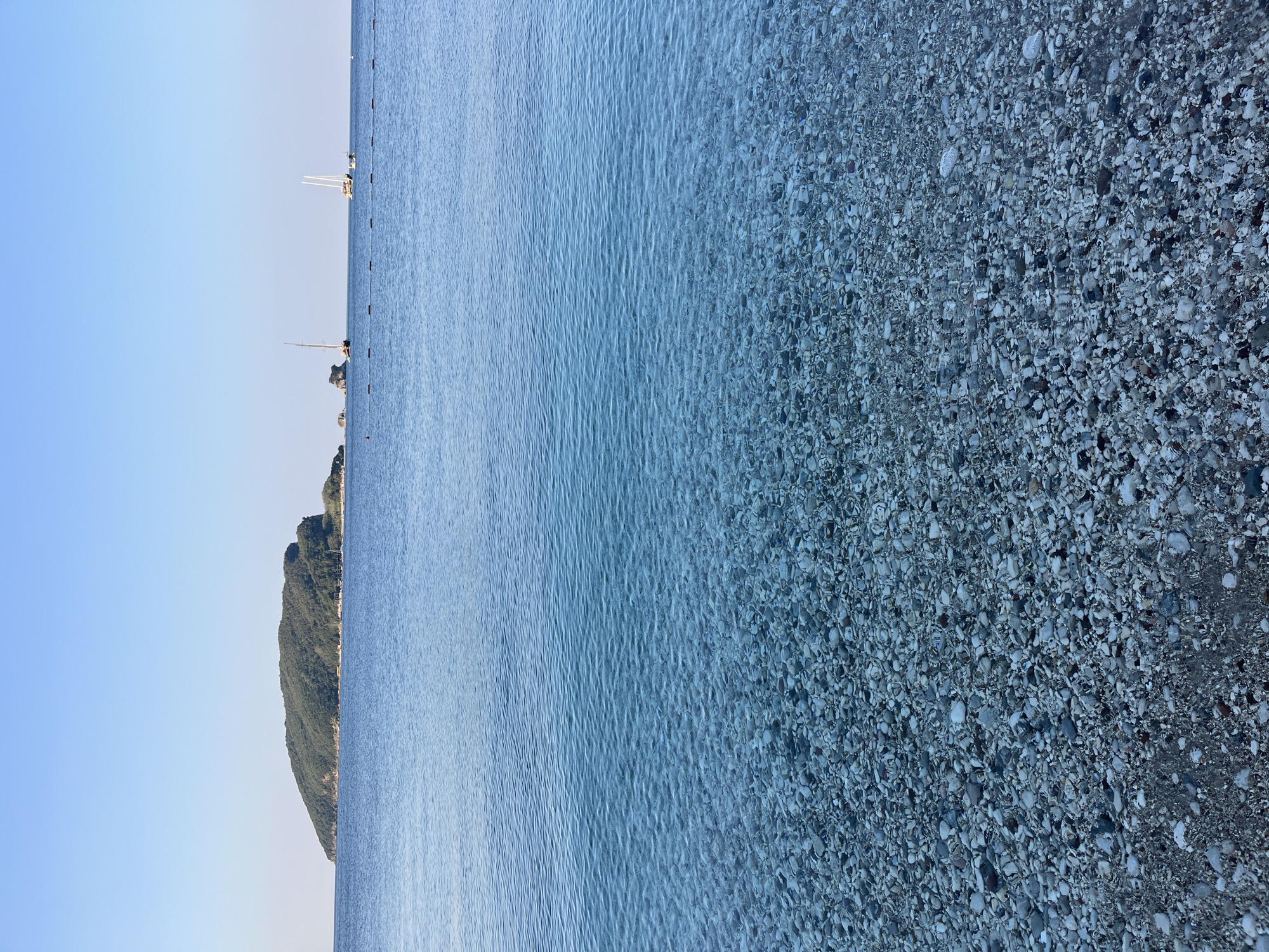 The image depicts a serene seascape with a clear, shallow sea revealing a rocky seabed in the foreground. The water is a vibrant turquoise, transitioning to a deeper blue towards the horizon. In the distance, a lush green island, identifiable as Sveti Nikola Island (also known as the "Montenegrin Hawaii"), dominates the left side of the frame. The island is covered in dense vegetation and slopes gently towards the sea. On the water, two sailboats with white sails are visible. The sky is a clear, pale blue, suggesting fair weather. The scene is likely a view from a beach in or near Budva, Montenegro, looking out towards Sveti Nikola Island.