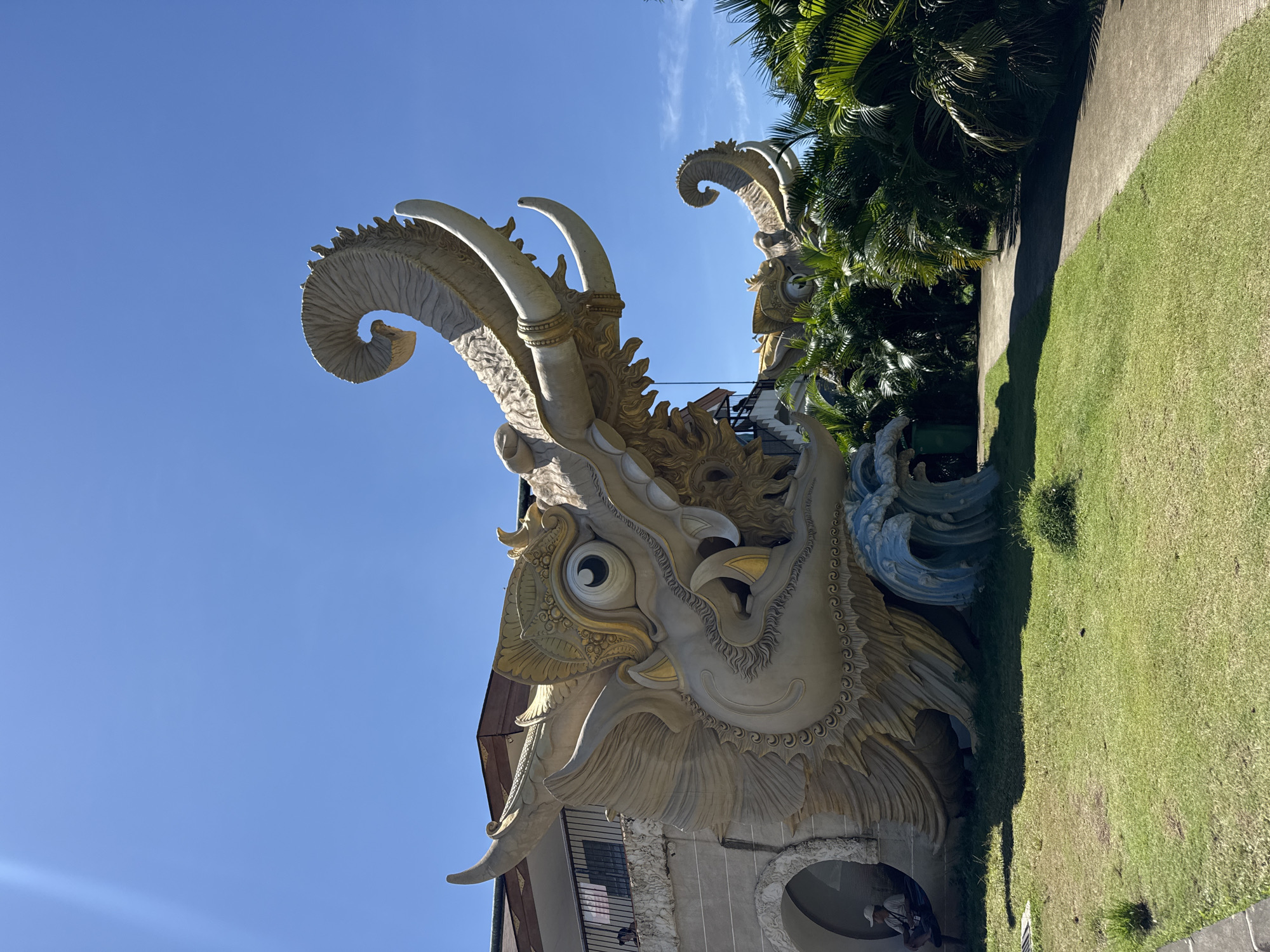 This image captures a monumental, highly ornate sculpture of a mythical creature, likely a dragon or a similar deity, forming a prominent part of a building's facade. The sculpture is primarily light-colored (off-white/cream) with intricate gold accents, particularly around its large, round eye and other detailed features. It boasts multiple large, curled horns or antlers, one prominently spiraling into the clear blue sky.
To the right of the main sculpture, another similar head is partially visible, nestled among dense, vibrant green palm trees. The setting is bright and sunny, with a clear blue sky overhead, suggesting a tropical environment. Below the statue, a neatly manicured lawn of green grass runs alongside a paved pathway. At the base of the main sculpture, near an arched entrance or opening, a person wearing a light-colored hat is partially visible. The overall scene conveys grandeur and intricate craftsmanship in an outdoor, sunny setting. The context points to a location in Denpasar, Indonesia. There is no visible text in the image.