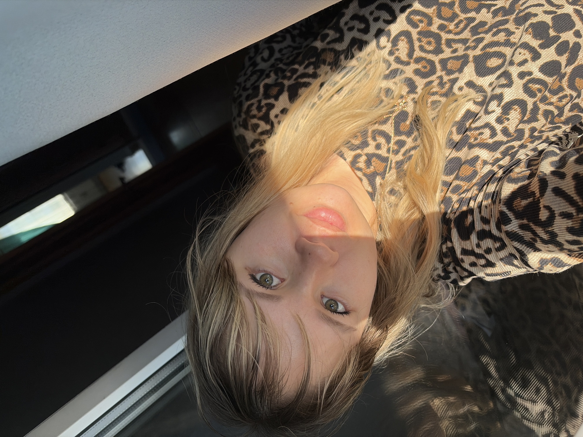 A fair-skinned person with light brown/blonde hair is positioned in an upside-down orientation relative to the frame. The individual's face, particularly the eyes, nose, and mouth, are illuminated, indicating a light source. The person is wearing a leopard-print top. The background shows a window frame and a dark, reflective surface.
There is no information available to determine that this image was taken in the city of Chaikovsky, Russia.