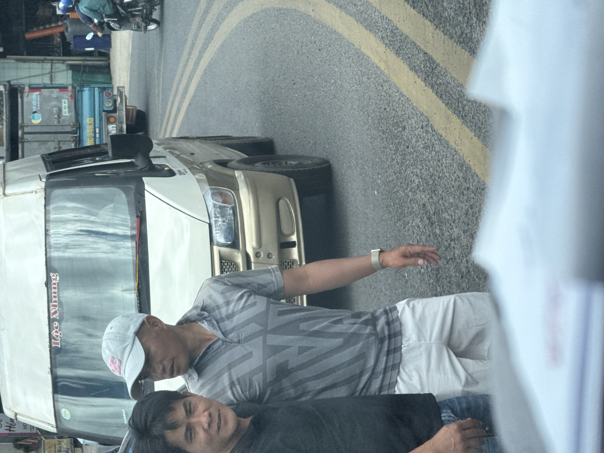 The image depicts a street scene in Phường Xuân Hương, Đà Lạt, Vietnam. In the foreground, two men are standing next to each other. The man on the left is wearing a black t-shirt and blue jeans. The man on the right is wearing a grey, patterned polo shirt and white shorts. He is also wearing a light grey baseball cap. They appear to be posing for a photo.
Behind them, a light-colored van is parked on the side of the road. The van has a sticker with red text that reads "Lộc Nhung". Further in the background, a light blue truck is visible, and a person on a motorcycle is riding away from the camera. The road has double yellow lines indicating the lane division. The environment suggests a warm climate, typical of Đà Lạt.