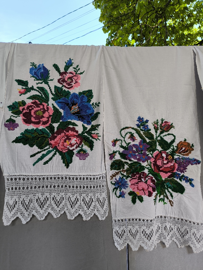 A full-frame, eye-level outdoor shot depicts two white, hand-embroidered textiles hanging vertically, likely on a clothesline. Each textile features intricate cross-stitch floral patterns in vibrant colors and a wide, white crocheted lace border at its lower edge. The textile on the left displays a large bouquet of pink, blue, and purple flowers with green foliage. The textile on the right exhibits a similar but distinct floral arrangement in pink, blue, purple, and orange hues. Both items appear to be hanging from an unseen support against a light gray vertical surface that occupies the lower half of the background. The upper half of the background reveals a clear blue sky, green tree foliage in the upper right, and faint utility lines across the sky. The scene is located outdoors in the city of Illintsi, Ukraine. No individuals are visible.