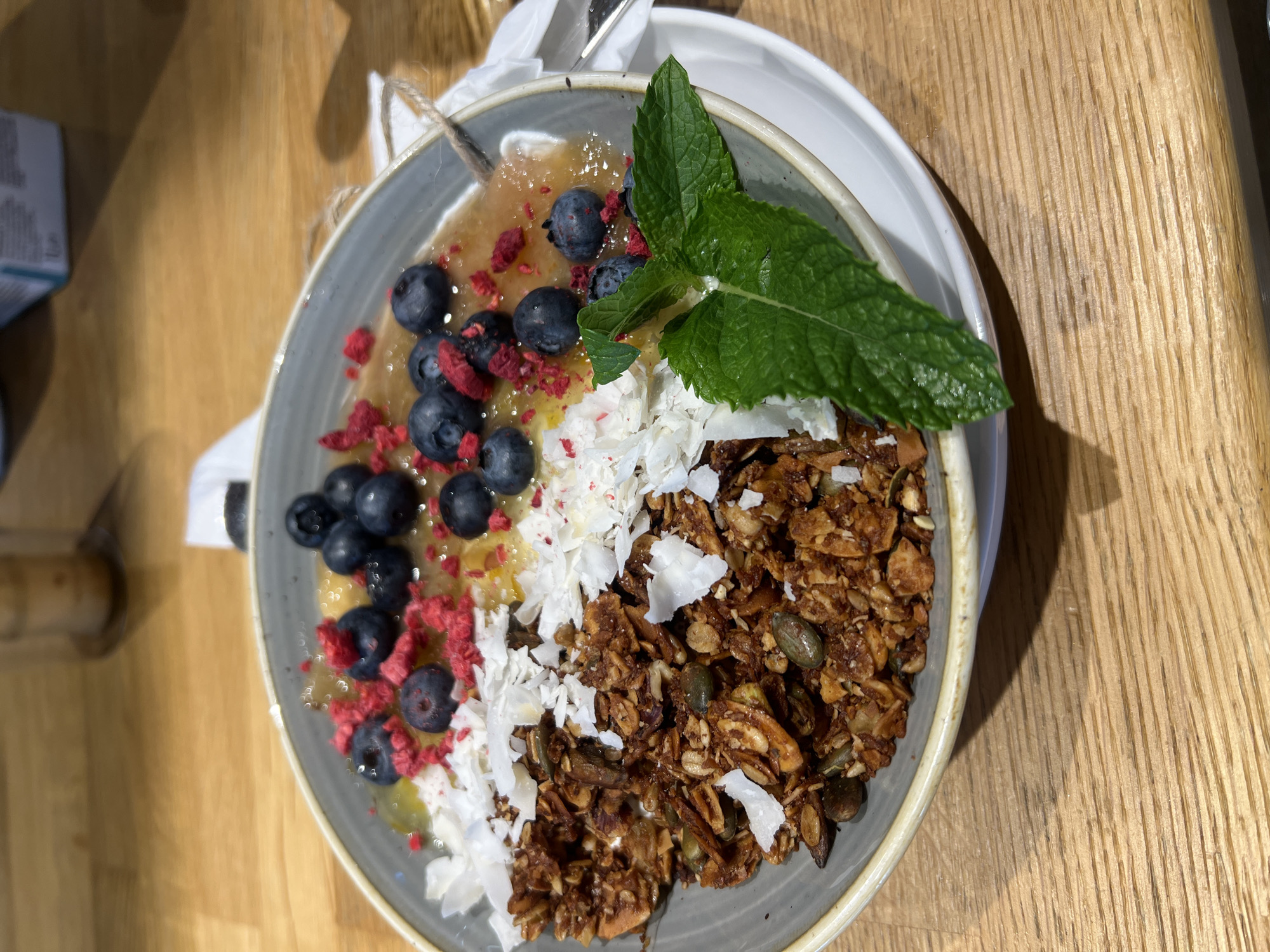 A close-up, eye-level shot shows a grey bowl filled with a healthy breakfast. The bowl contains granola with visible seeds and coconut flakes, a fruit puree, and is topped with blueberries and freeze-dried raspberries. A sprig of mint garnishes the dish. The bowl sits on a white saucer on a wooden table, suggesting a cafe or restaurant setting. The image is sharp and well-lit, highlighting the textures and colors of the food. The location is the city of Vysoké Tatry, Slovakia.