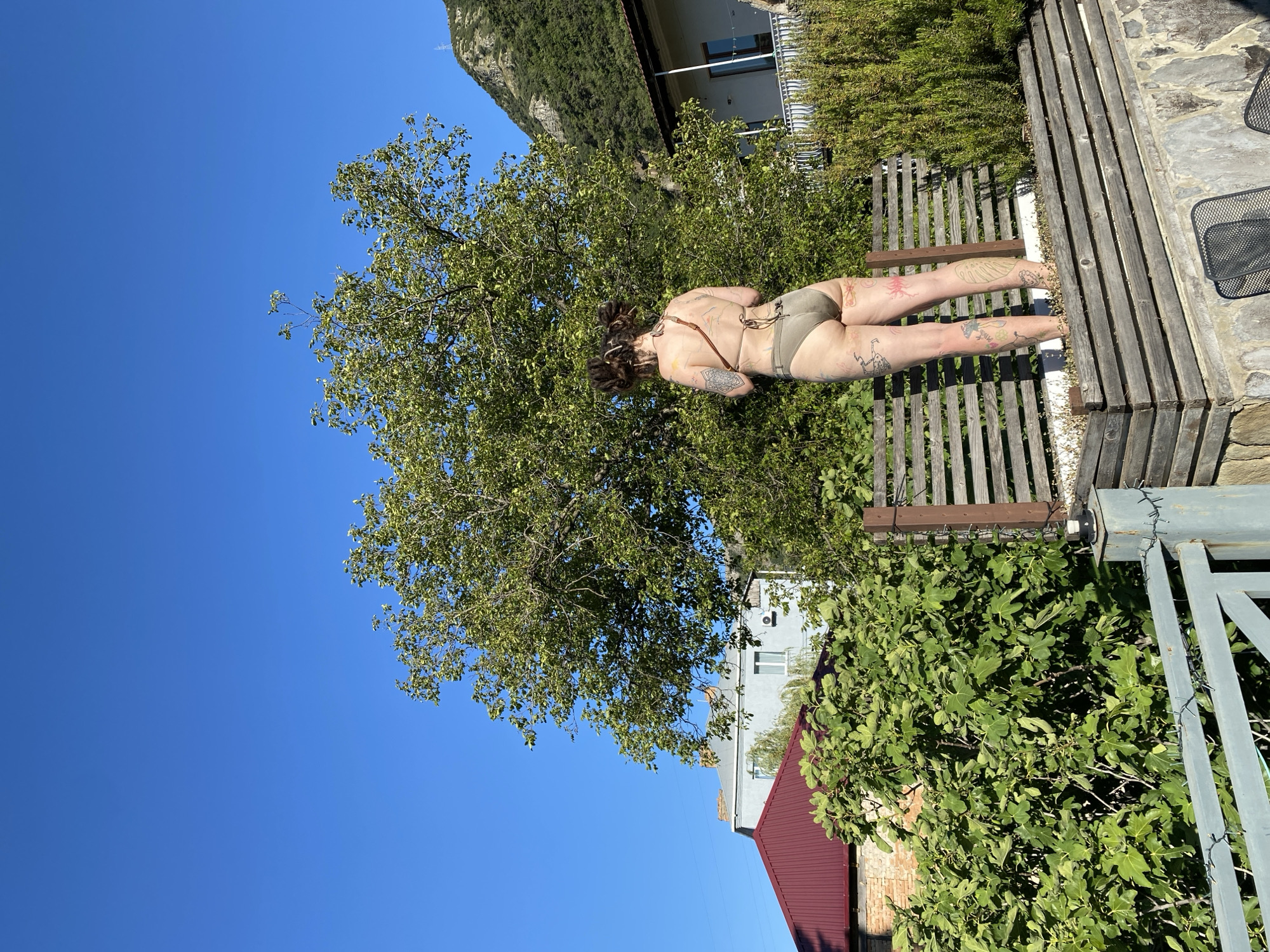 A person with multiple tattoos is standing outdoors, facing away from the camera, in what appears to be a backyard or garden area. They are wearing a bikini. The person has their arms behind their back and their head tilted slightly. The scene includes lush green foliage from trees and bushes, a wooden fence, and a stone wall. In the background, there is a building with a white facade and windows, and a red-tiled roof. A steep, forested mountain is visible in the distance. The sky is a clear, vibrant blue, suggesting a sunny day, likely late morning or early afternoon. The overall impression is one of relaxation and enjoying the outdoors. No visible text is present in the image.