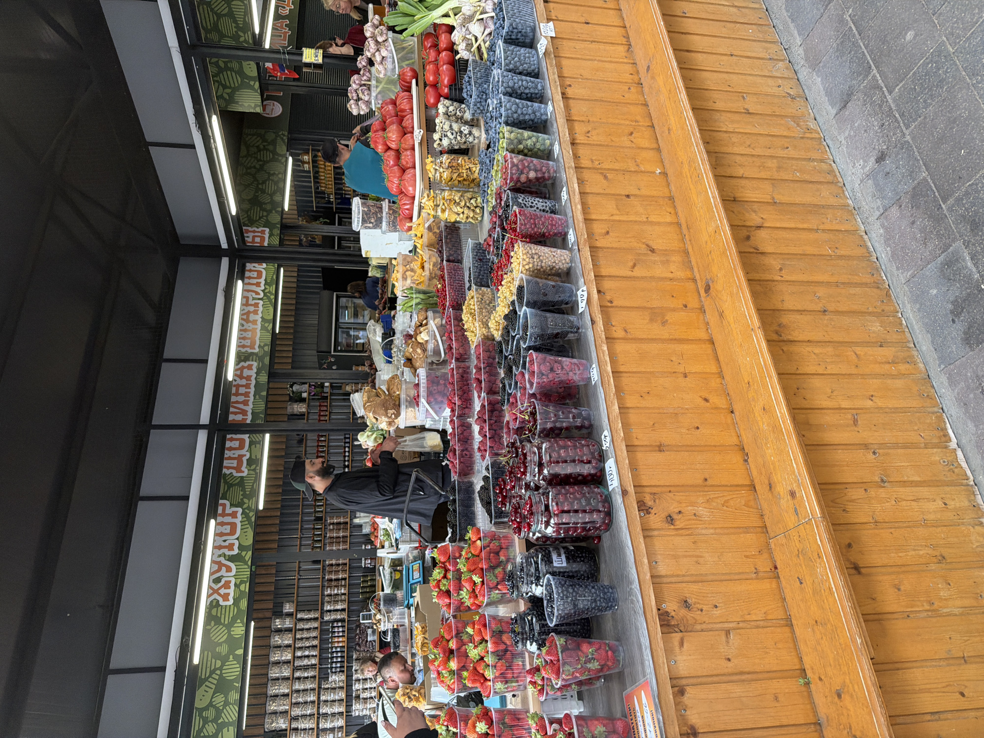 The image depicts a stall at an outdoor market in Kaliningrad, Russia, likely selling fresh produce. In the foreground, a wooden counter displays an abundance of assorted berries and fruits in various containers. These include strawberries, blueberries, raspberries, blackberries, and cherries, some in plastic cups and others in larger jars. Behind the produce, a man wearing a black cap and shirt stands near a scale, seemingly attending to a customer or restocking. Other individuals are visible in the background, including shoppers and stall workers. The stall is covered by a dark awning, and shelves laden with jars and containers are visible in the background. The overall atmosphere suggests a vibrant and busy marketplace.