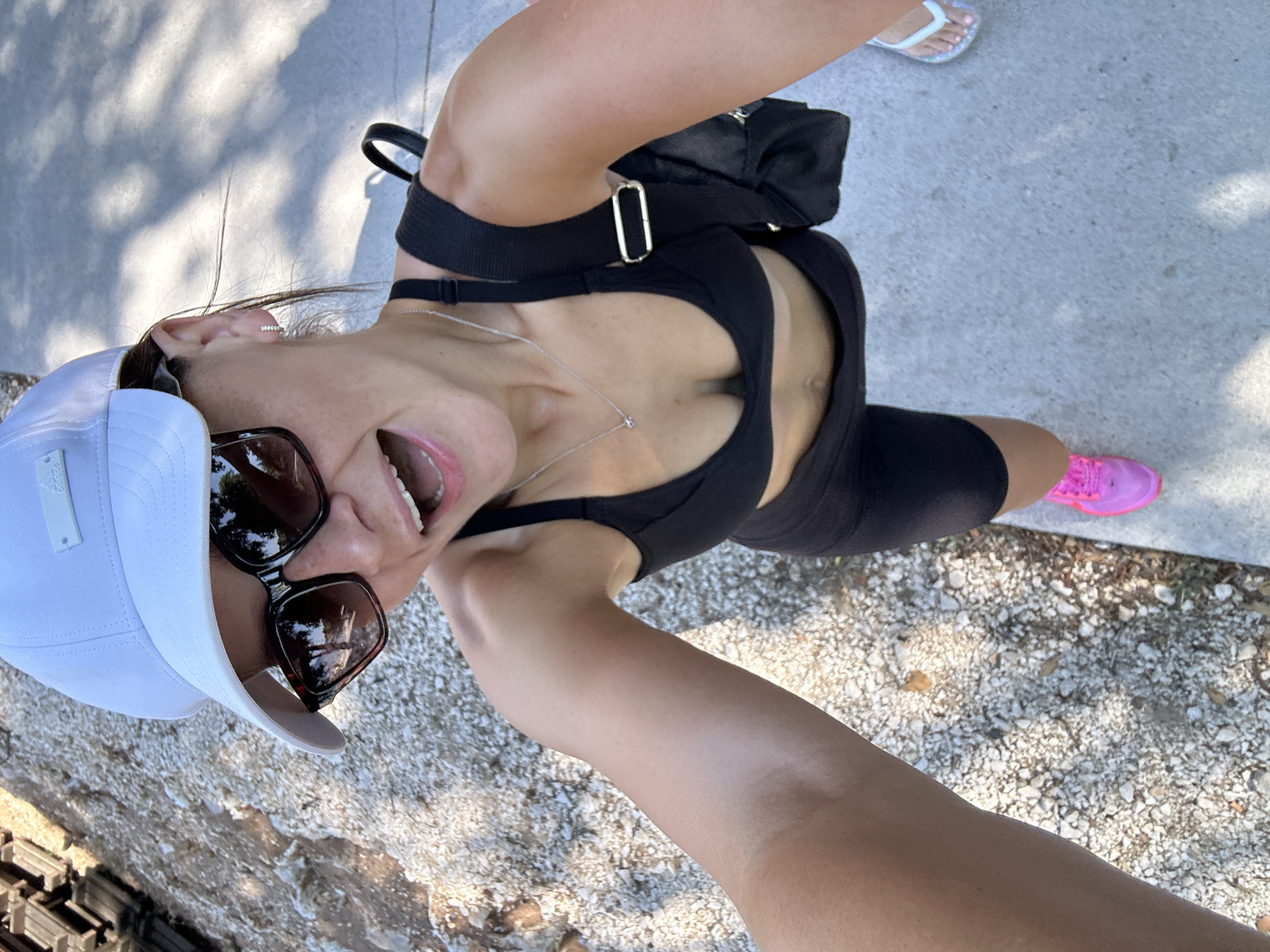 The image shows a woman taking a selfie outdoors, wearing a white baseball cap, sunglasses, a black sports bra, and black shorts. She has bright pink running shoes on her feet, and a small backpack is visible on her back. She appears to be smiling or laughing with her mouth open. The background consists of a gravel path on the left and a concrete path on the right, with dappled sunlight creating shadows. The setting appears to be a casual outdoor environment, possibly a park or a recreational area.