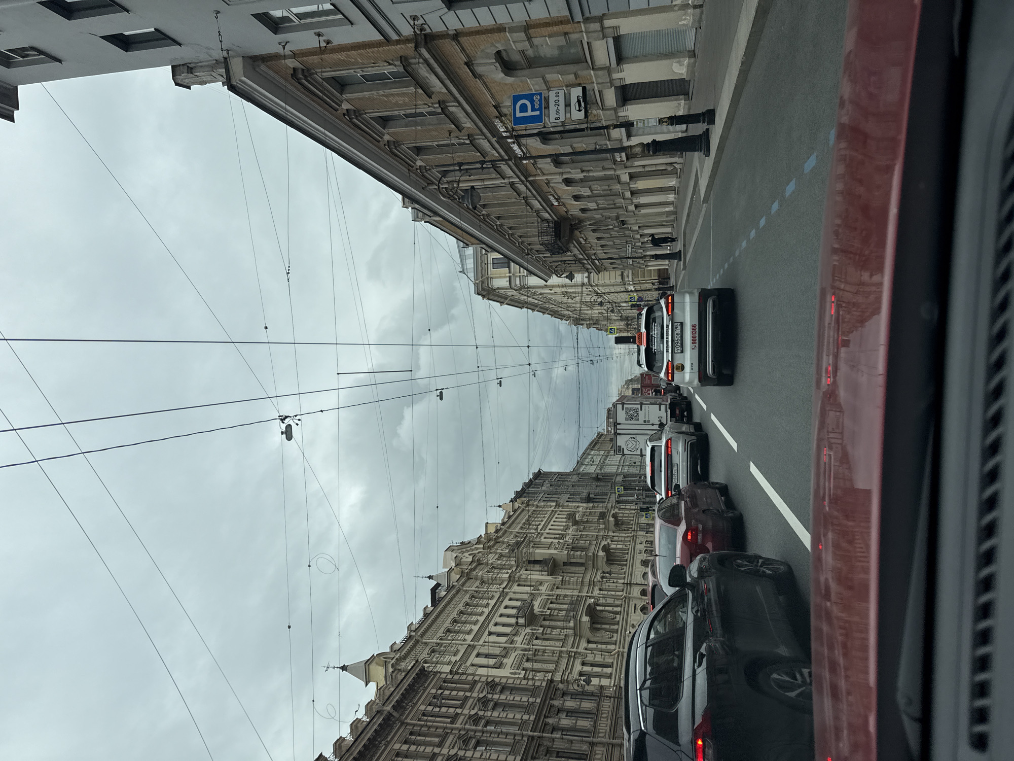 The image depicts a street scene in an urban area. The street is lined with buildings of elaborate architectural style. Overhanging wires stretch across the sky above the street. A parking sign is visible on one of the buildings. Cars are lined up in traffic on the street. The image appears to be taken from within a vehicle, with a portion of the vehicle's interior visible on the right side of the frame.