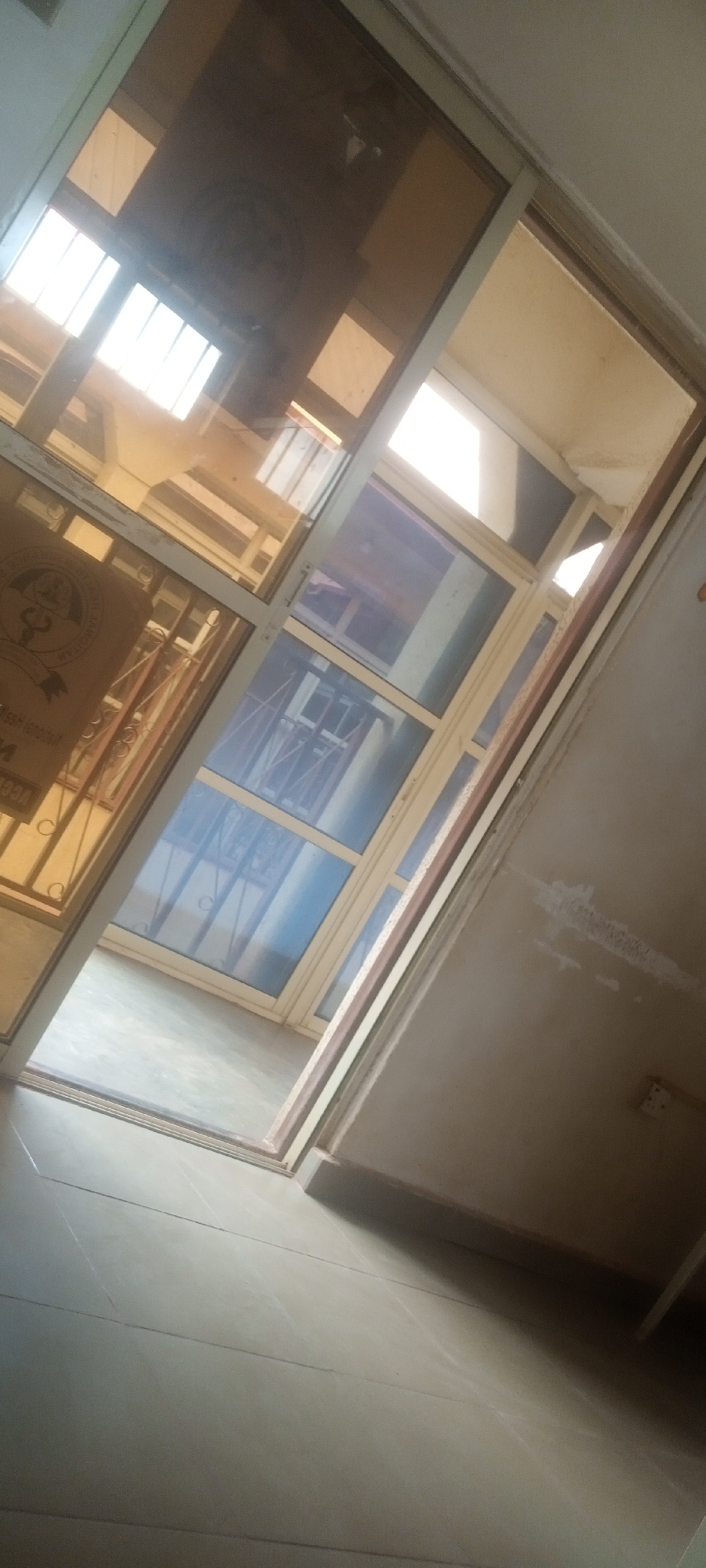 The image shows a view through a glass door, looking towards a room with blue-tinted windows and a metal security gate in the background. The glass door is partially open, and a logo or emblem is visible on the left side of the glass. The floor in the foreground is tiled. The wall to the right of the door is beige and has an electrical outlet. The location is Yelwa, Nigeria.