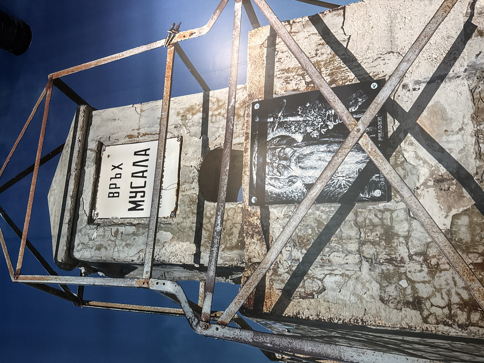 The image shows a low-angle view of a concrete structure with a metal frame. A sign reads "ВРЪХ МУСАЛА" in Cyrillic. A black and white photograph is attached to the structure. The background is a blue sky. This structure is at the peak of Musala, the highest peak in the Rila Mountains in Bulgaria, which are located near the city of Sofia.
