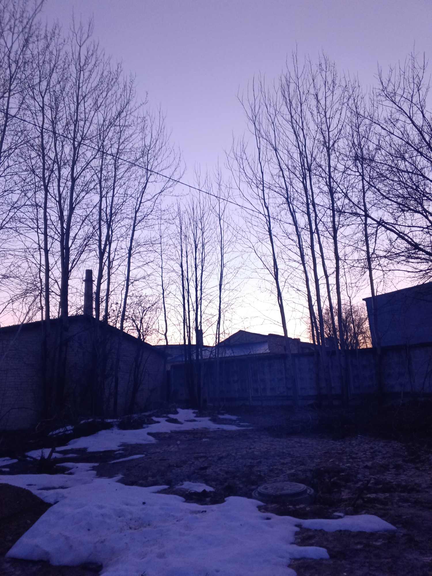 The image depicts an outdoor scene in Красный Маяк, Russia, at dusk. The foreground is dominated by patches of melting snow on uneven ground, with a manhole cover visible near the center. A low fence and the outlines of several low buildings can be seen in the middle ground, partially obscured by tall, leafless trees. In the background, the sky is a gradient of purple and pink, suggesting either sunrise or sunset. There are no people visible in the image.