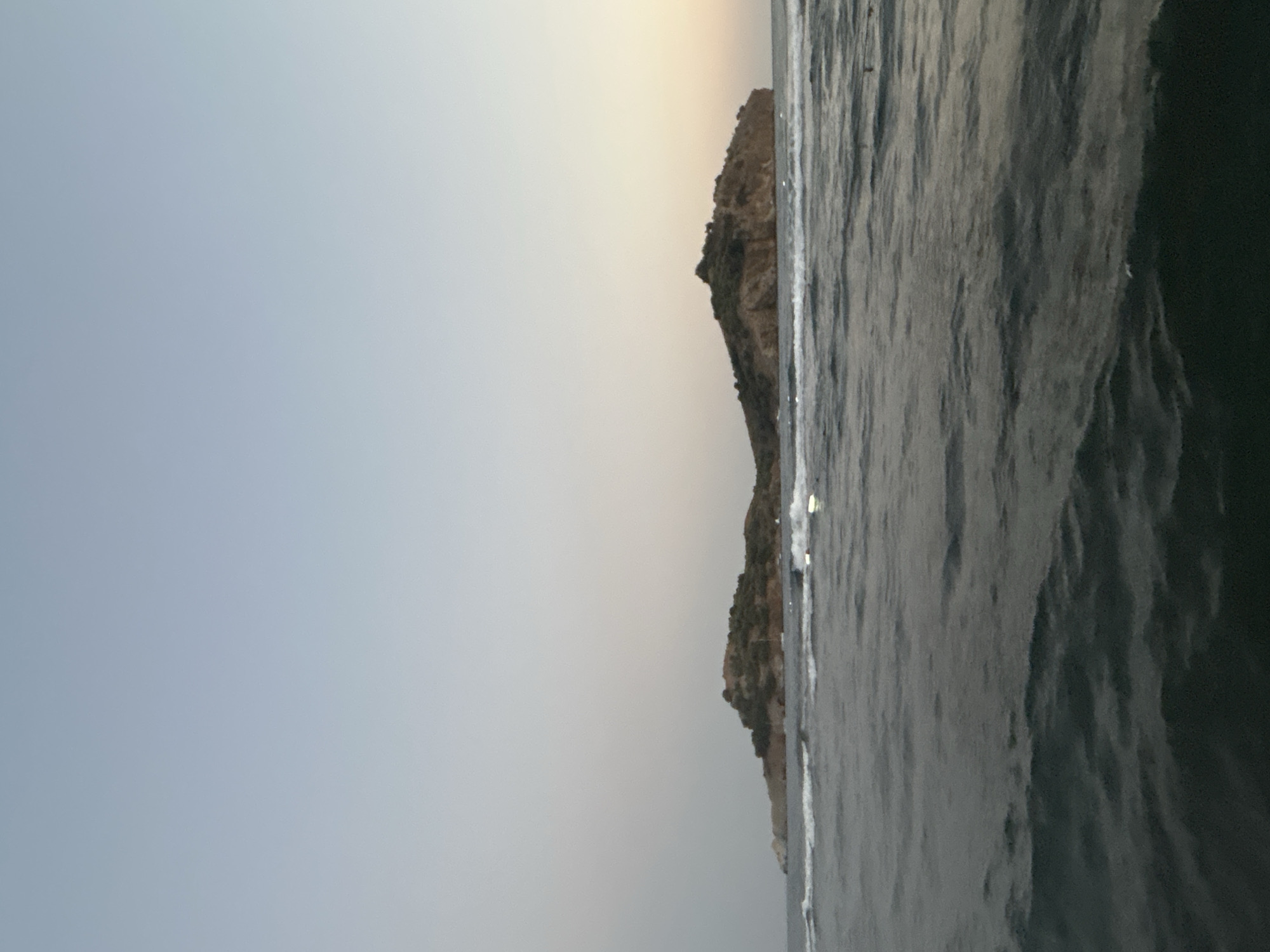 An island with scattered buildings is visible in the distance, situated in the sea. The island is covered with green vegetation and rocky terrain. The water in the foreground is dark and choppy, with small waves. The sky above is a gradient of light blue and gray, suggesting it may be early morning or late evening. The overall scene is a natural landscape with a hint of human presence on the island. The location is the sea off the coast of Ortaca, Turkey.