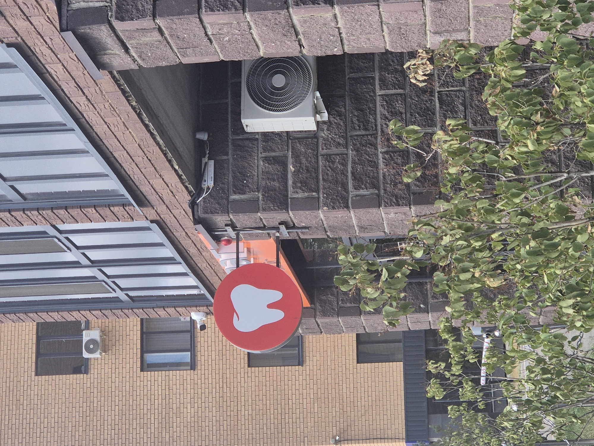 The image shows a close-up of a brick building facade with a red circular sign featuring a white tooth graphic, indicating a dental clinic. An outdoor air conditioning unit is mounted on the brick wall to the right of the sign. In the background, a lighter-colored brick building with multiple windows is visible. There are also security cameras mounted on the building. The scene appears to be outdoors, possibly on a street or walkway, with some greenery in the foreground obscuring the lower part of the buildings. The environment suggests an urban setting.