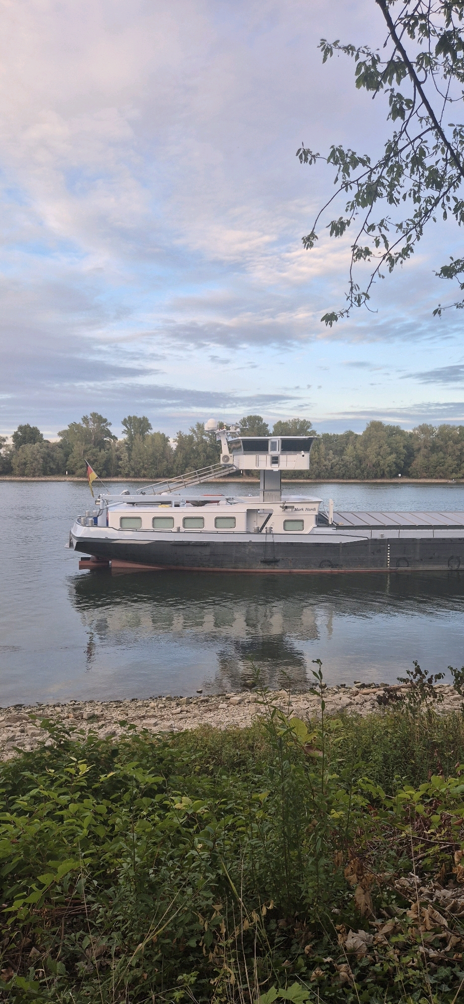 An aerial view taken from a high vantage point overlooking a body of water with a barge docked near the bank. The barge is a long, dark vessel with a white superstructure. A German flag is visible on the left side of the barge. The water is calm, with a reflection of the barge visible on its surface. The shore is lined with rocks and vegetation. In the background, there is a dense line of trees. The sky is partly cloudy with a soft, diffused light, suggesting it may be early morning or late afternoon. The scene appears to be located on the Rhine River near Mainz, Germany.