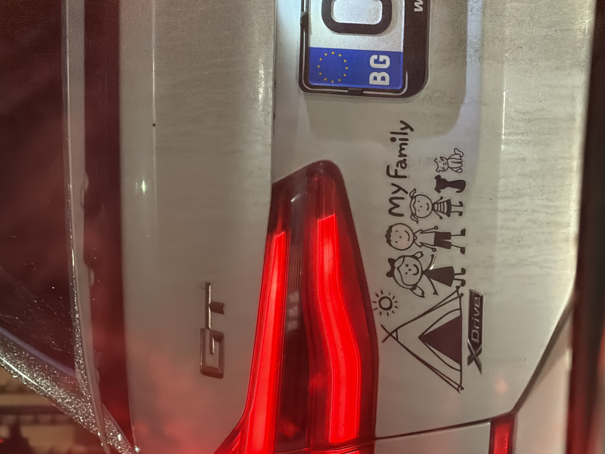 The image shows the rear of a silver BMW GT car. The car has a Bulgarian license plate with the "BG" country code. A "My Family" sticker with stick figure representations of a family and animals is visible, along with a drawing of a tent and the "XDrive" emblem. The "GT" badge is affixed to the body. The car's tail lights are illuminated, suggesting it's nighttime or low-light conditions. The overall scene suggests a car in Bulgaria. No people are directly visible. The location is not explicitly confirmed as Sofia, but based on the Bulgarian license plate, it's likely within the country.