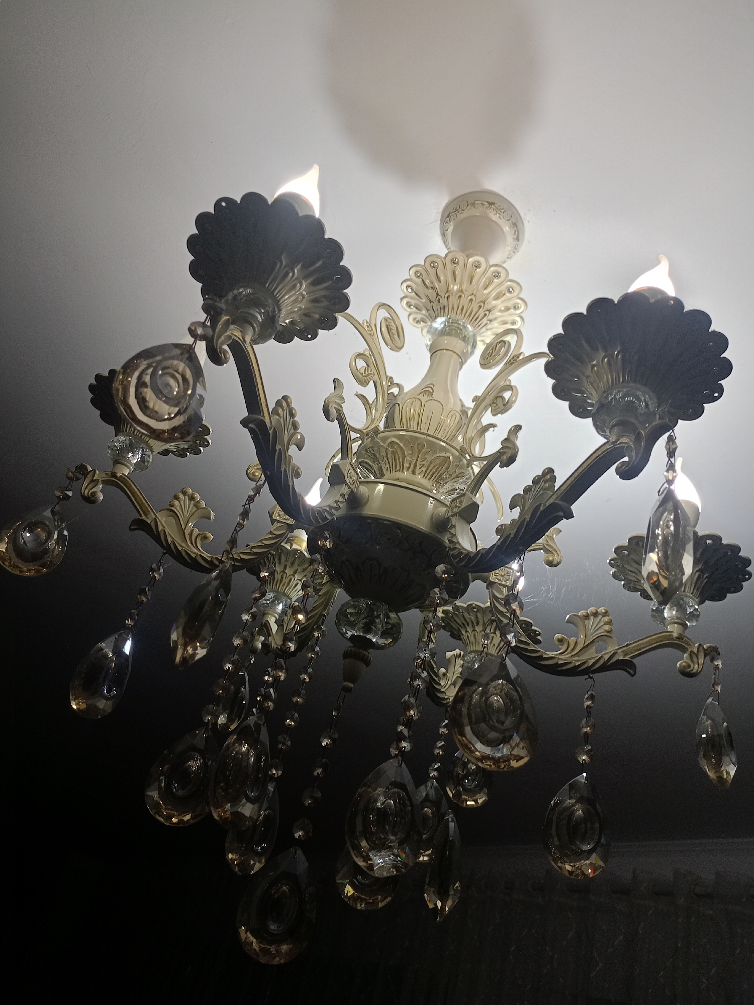A vintage-style chandelier hangs from a white ceiling in a dimly lit room. The chandelier is elaborate, with multiple arms supporting decorative elements and faux candle lights. It is adorned with numerous faceted crystal droplets that refract the light, creating a sparkling effect. The metal components of the chandelier appear to be brass or a similar antique-finish metal, with some ornate detailing. There are also what appear to be cobwebs present on some of the chandelier's arms, suggesting it might not be recently cleaned. The background is dark, indicating low ambient light apart from the chandelier itself. The image is taken from a low angle, looking up at the chandelier. There is no information about the city of Kandauraul, Russia, present in the image.