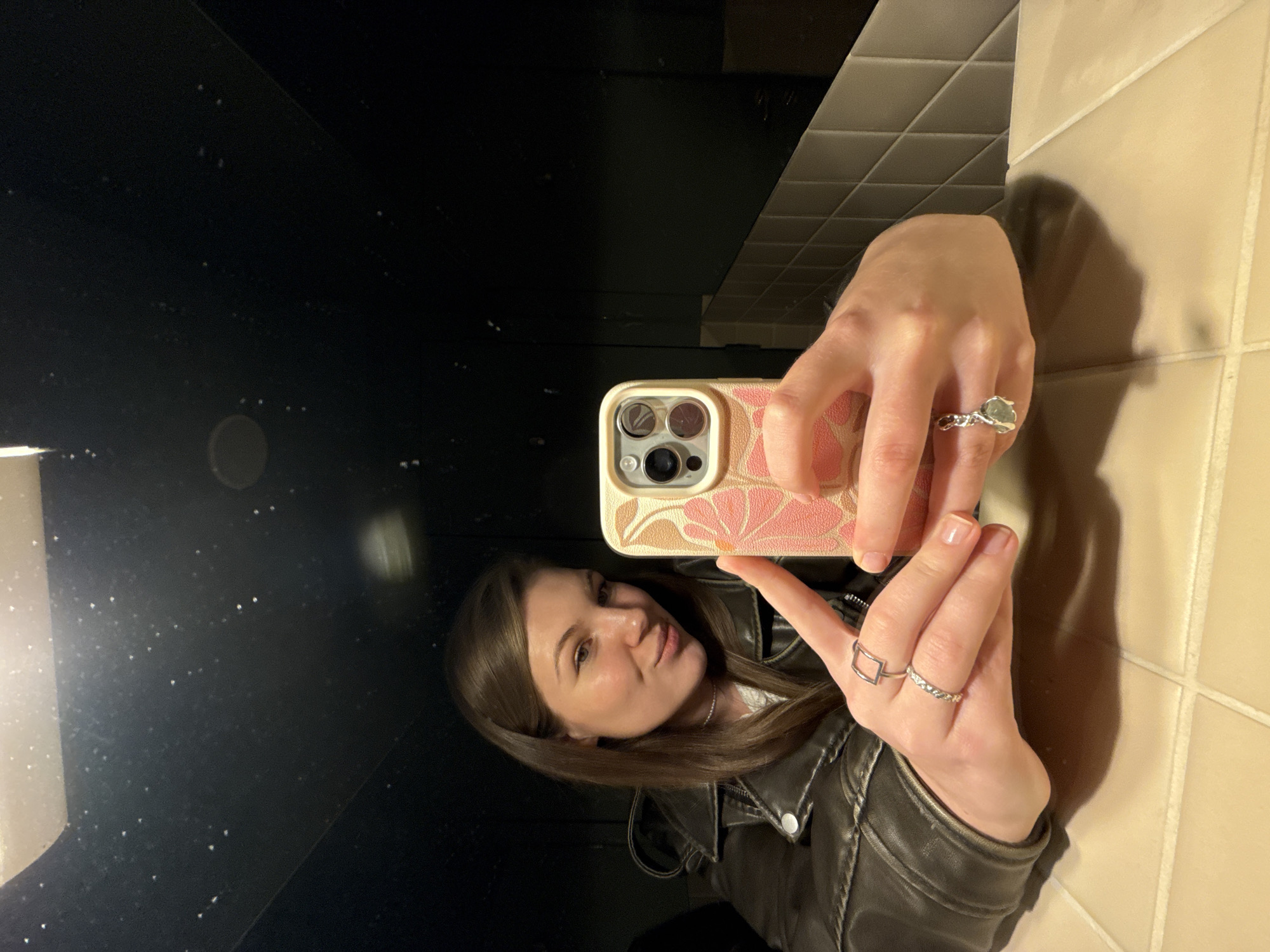 A person is taking a selfie in a bathroom mirror. The person is wearing a brown leather jacket and rings on both hands. The phone case is patterned with pink and beige floral designs. The mirror reflects a dark space. The bathroom features beige tiled walls. It is not possible to determine the location in Belgrade, Serbia, from the visual information provided.