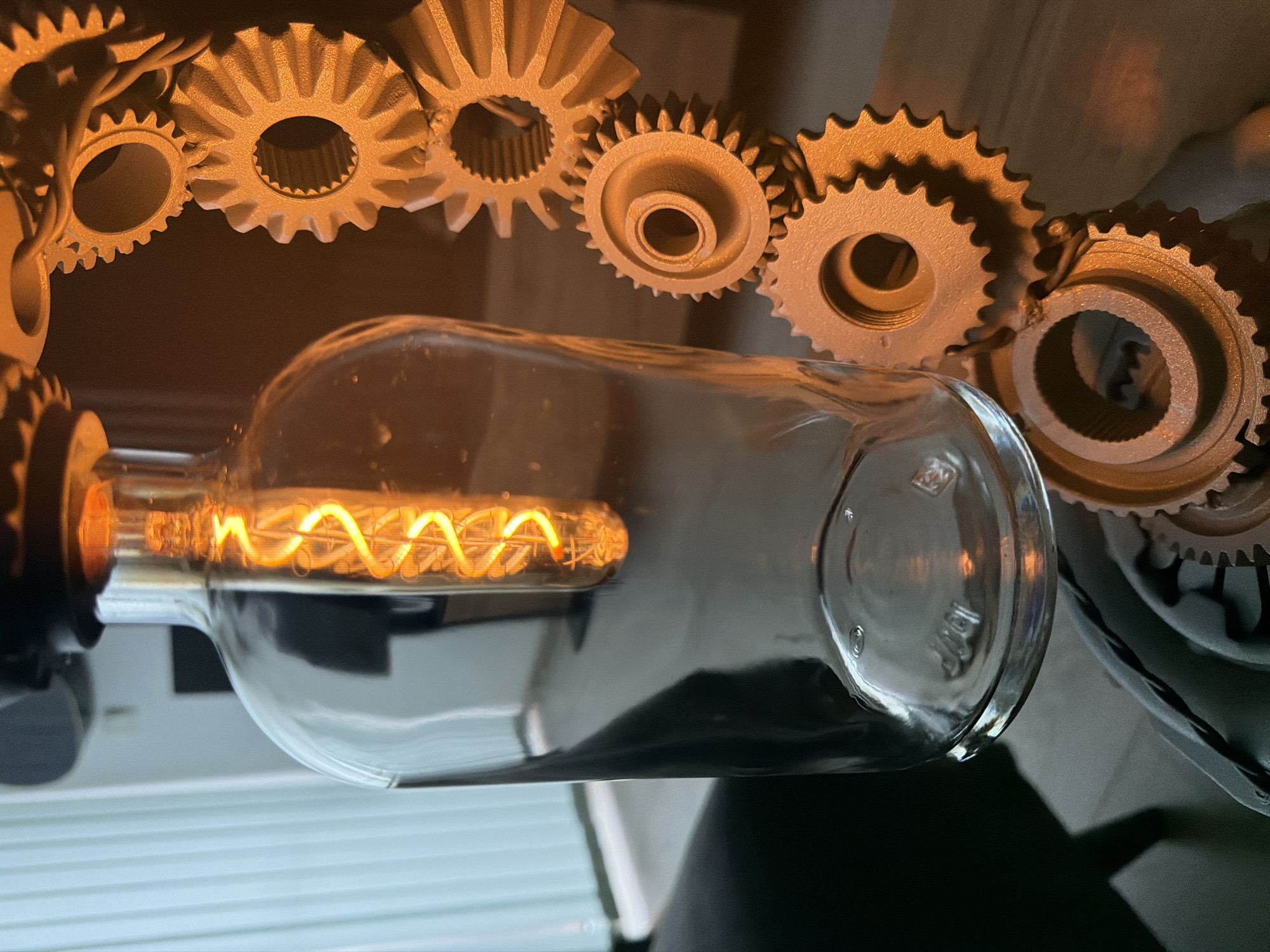 This horizontal image, captured in Sofia, Bulgaria, features a unique, steampunk-inspired decorative lamp. The central element is a clear glass bottle, positioned horizontally, containing an illuminated light bulb with a prominent coiled filament that emits a warm, bright orange-yellow glow.
Arranged around the bottle are several gear-like components of various sizes and designs. These objects appear to be made of a metallic or metal-painted material, possibly bronze or gold, and exhibit textured surfaces and mechanical details such as central splines and teeth. They form an intricate, decorative frame or base for the lamp.
The setting is an indoor space, characterized by a dark, indistinct background. Below the light fixture, glimpses of a light-colored, ribbed surface, possibly blinds or a radiator, are visible, alongside darker areas suggesting furniture or other room elements. The illumination of the bulb indicates the lamp is turned on, casting a cozy ambiance. The artificial light suggests it is either evening or the room is dimly lit. No specific weather conditions are discernible.
Faint, embossed details, possibly manufacturing codes or volume indicators such as "130" and a symbol resembling a diamond, are visible on the clear glass bottle, particularly near its base in the cropped sections, though they are partially obscured by reflections.