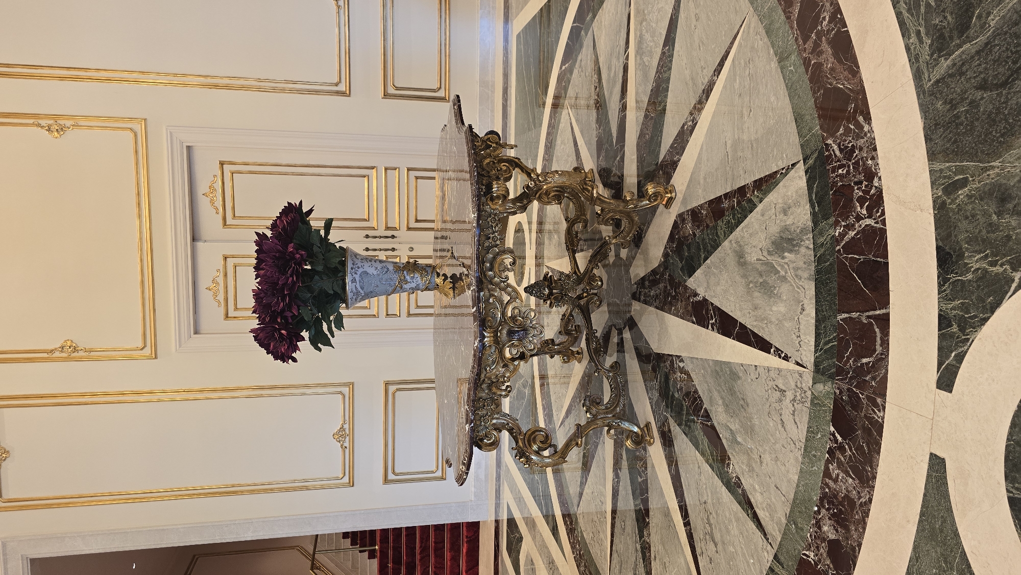 The image shows an ornate table with a glass top and a golden, scrolled base. A vase filled with deep purple flowers sits on the table, which is positioned on a marble floor with a geometric pattern of green, white, and dark red tiles. The background features a white wall with elaborate gold trim molding, creating framed panels. The overall aesthetic suggests a luxurious interior space. The orientation of the image is such that the floor is at the bottom and the wall is at the top, with the table placed horizontally across the center. There is a glimpse of a red carpeted staircase at the bottom left of the frame. The lighting appears to be indoor artificial light, possibly from overhead fixtures, casting soft shadows and highlighting the metallic sheen of the table's base. The scene is static and does not depict any activity or people. The setting evokes a sense of opulence and grandeur, possibly in a hotel lobby, a grand hall, or a stately home. The location context provided, Agios Epiktitos, Northern Cyprus, hints at a Mediterranean setting, but this is not visually confirmed within the image itself. No text is visible in the image.