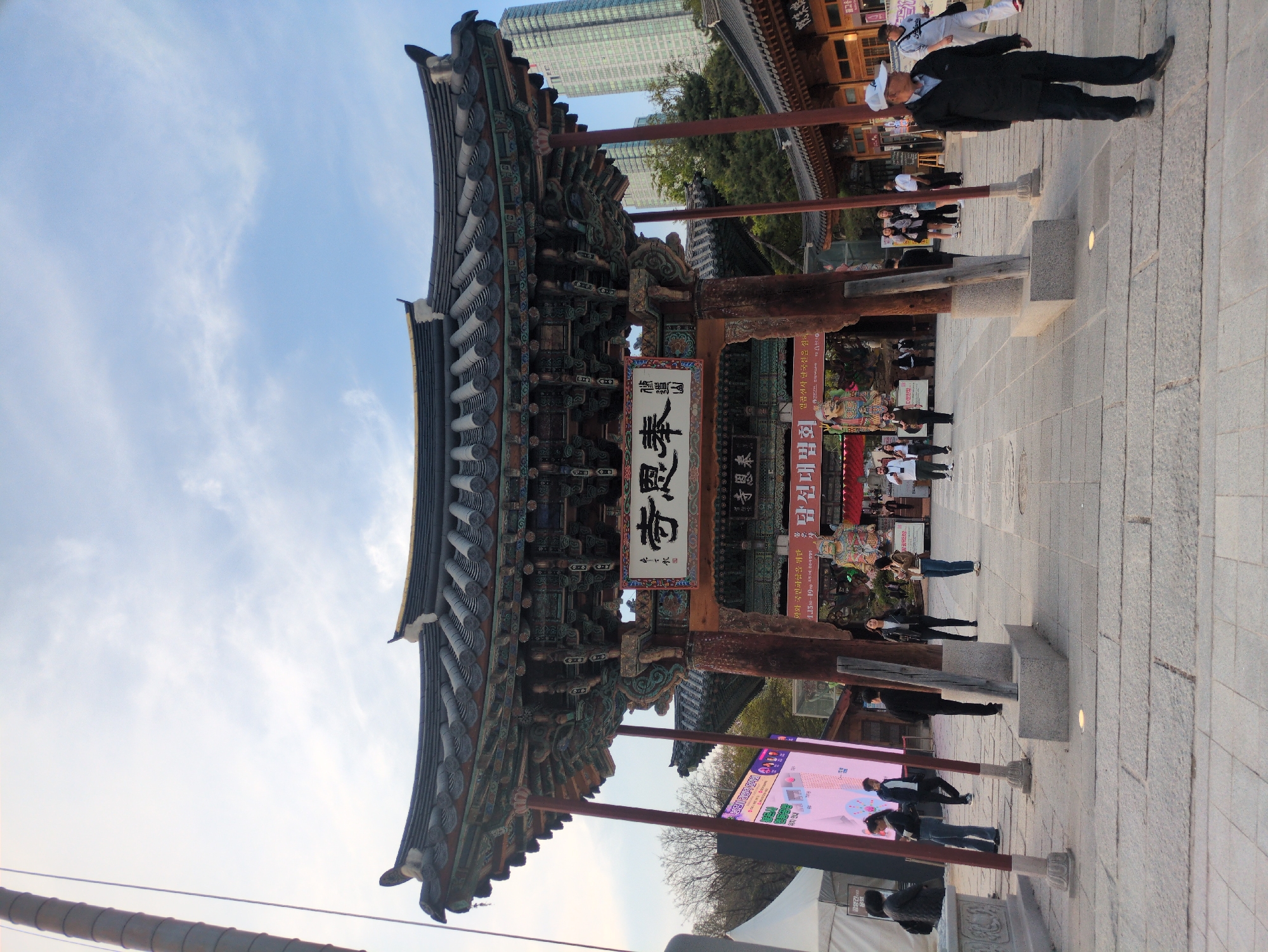 This image shows the entrance to Bonguensa Buddhist Temple in Seoul, South Korea. The traditional Korean-style gate is made of wood with intricate carvings and a tiled roof. A sign with Korean characters hangs from the gate. People are walking around the temple grounds, and a modern building can be seen in the background. A signboard shows information on a screen.