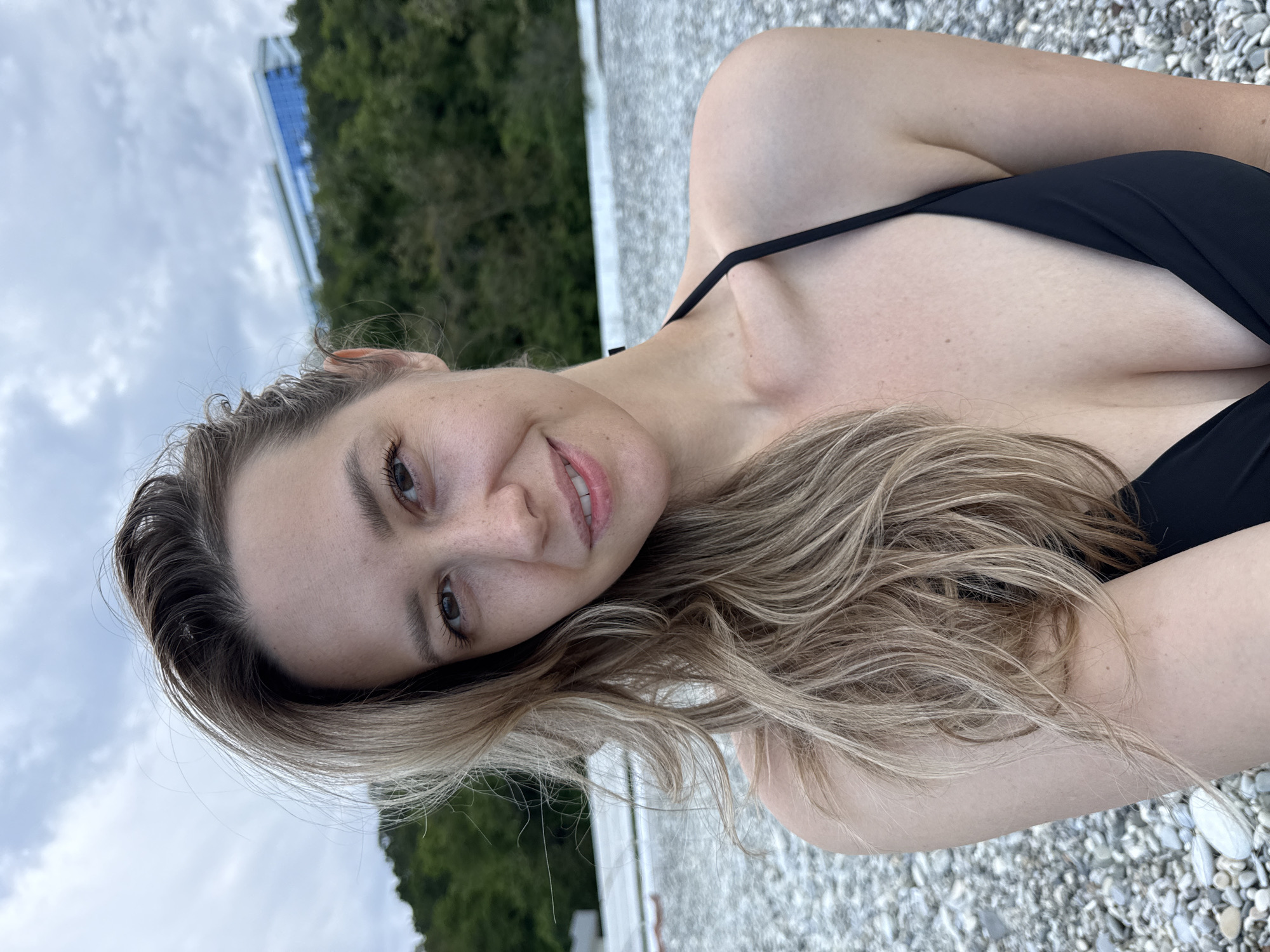 The image is a medium shot selfie of a woman with blonde hair, wearing a black bikini, on a pebble beach. She is looking at the camera and smiling. In the background, there is a body of water, a hill with trees, and a modern building. The location is identified as the city of Nebug, Russia. The sky is overcast.