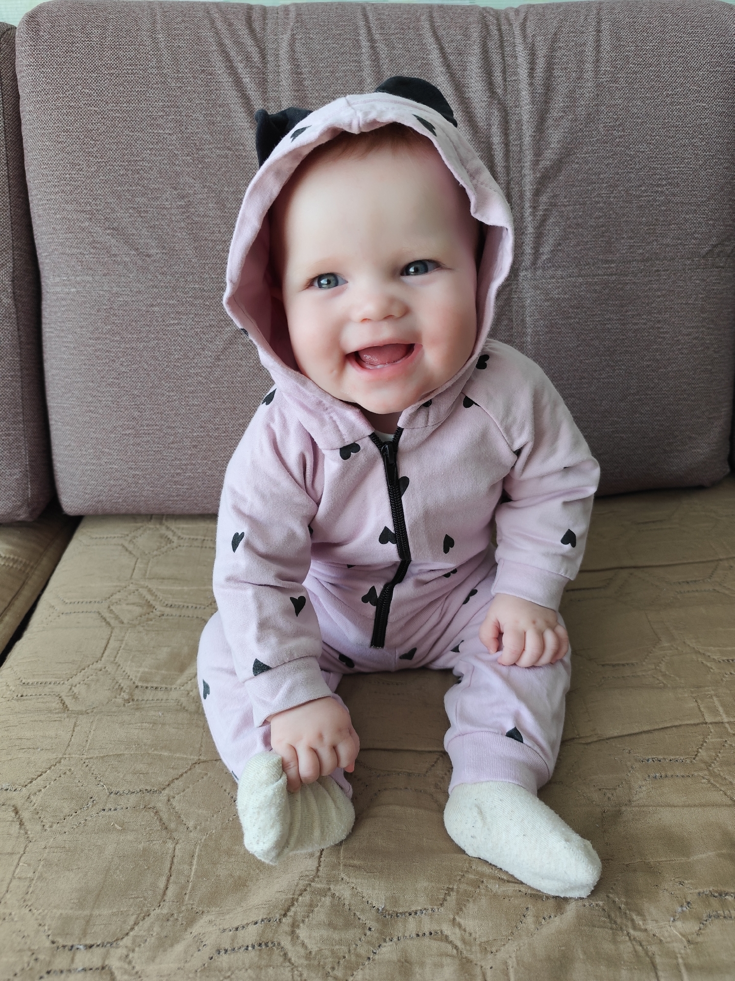 A baby, dressed in a pink onesie with small black hearts and a hood with black ears, is sitting on a brown couch with a pillow behind. The baby is smiling and looking directly at the camera, with their mouth open and tongue visible. The baby is holding their foot with one hand. The location is indoors, likely a home, and is not visibly in Фоки, Russia.