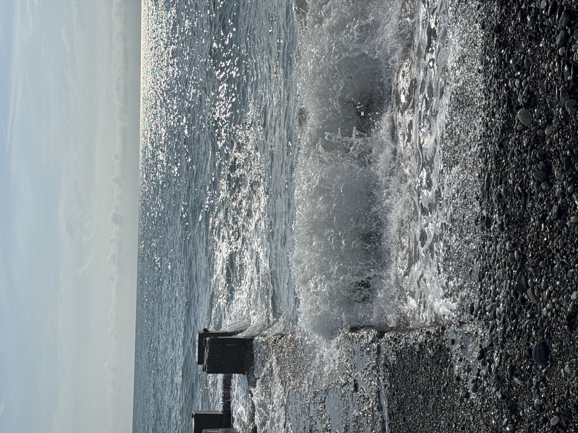 The image depicts a seascape, likely taken at a beach in Sochi, Russia. The scene includes a rocky beach with small pebbles, waves crashing against a concrete structure or pier, and the sea stretching to the horizon under a bright sky. The water is shimmering with sunlight, and the wave action creates foam and spray. There are no visible people in the image. The photograph appears to have been taken during daylight hours.
